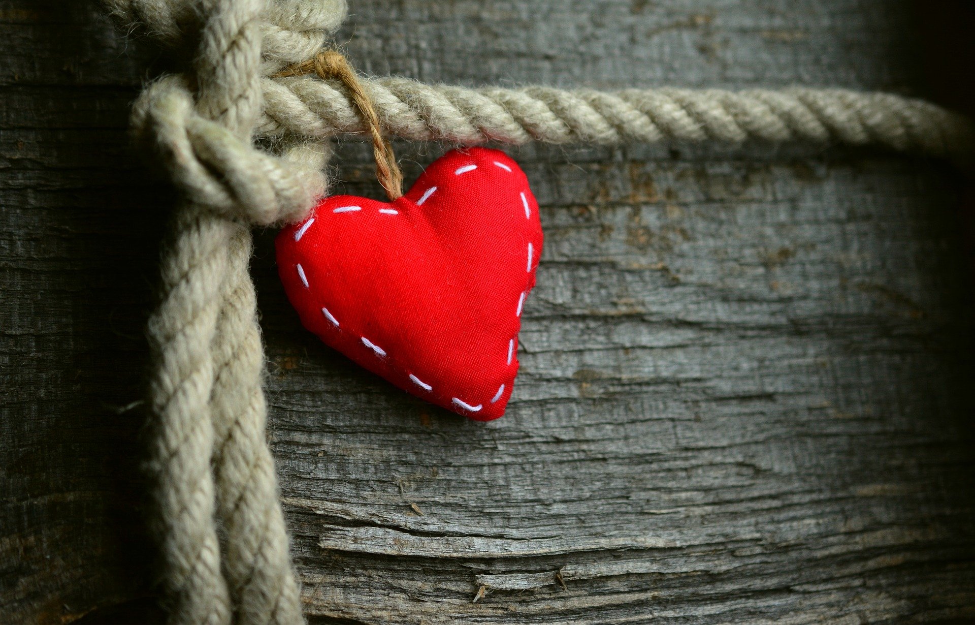 A red fabric heart with white stitches hanging from a beige rope on a weathered wooden surface.