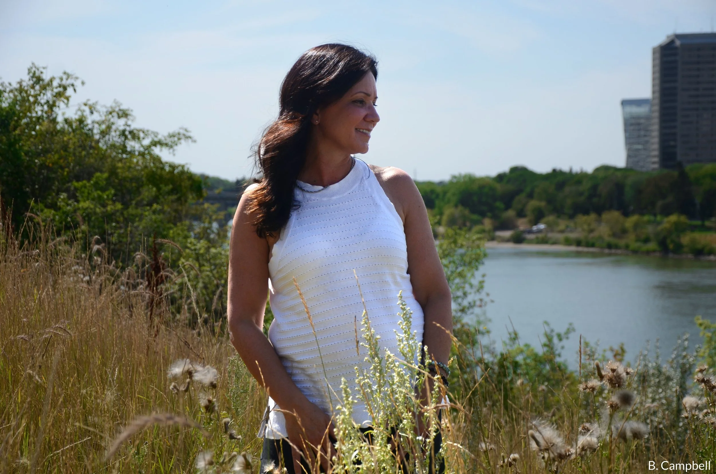Woman with dark hair wearing a white sleeveless top standing in tall grass near a river with trees and high-rise buildings in the background on a sunny day.