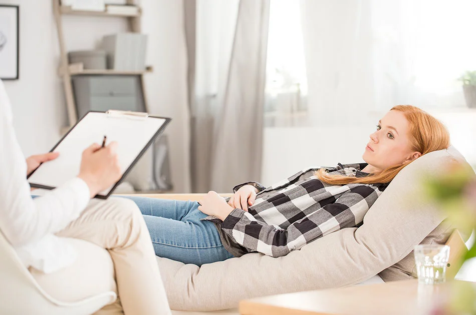 A young woman with red hair lying on a cushioned chair during a therapy or counseling session, listening to a therapist holding a clipboard and taking notes in a bright, cozy room.
