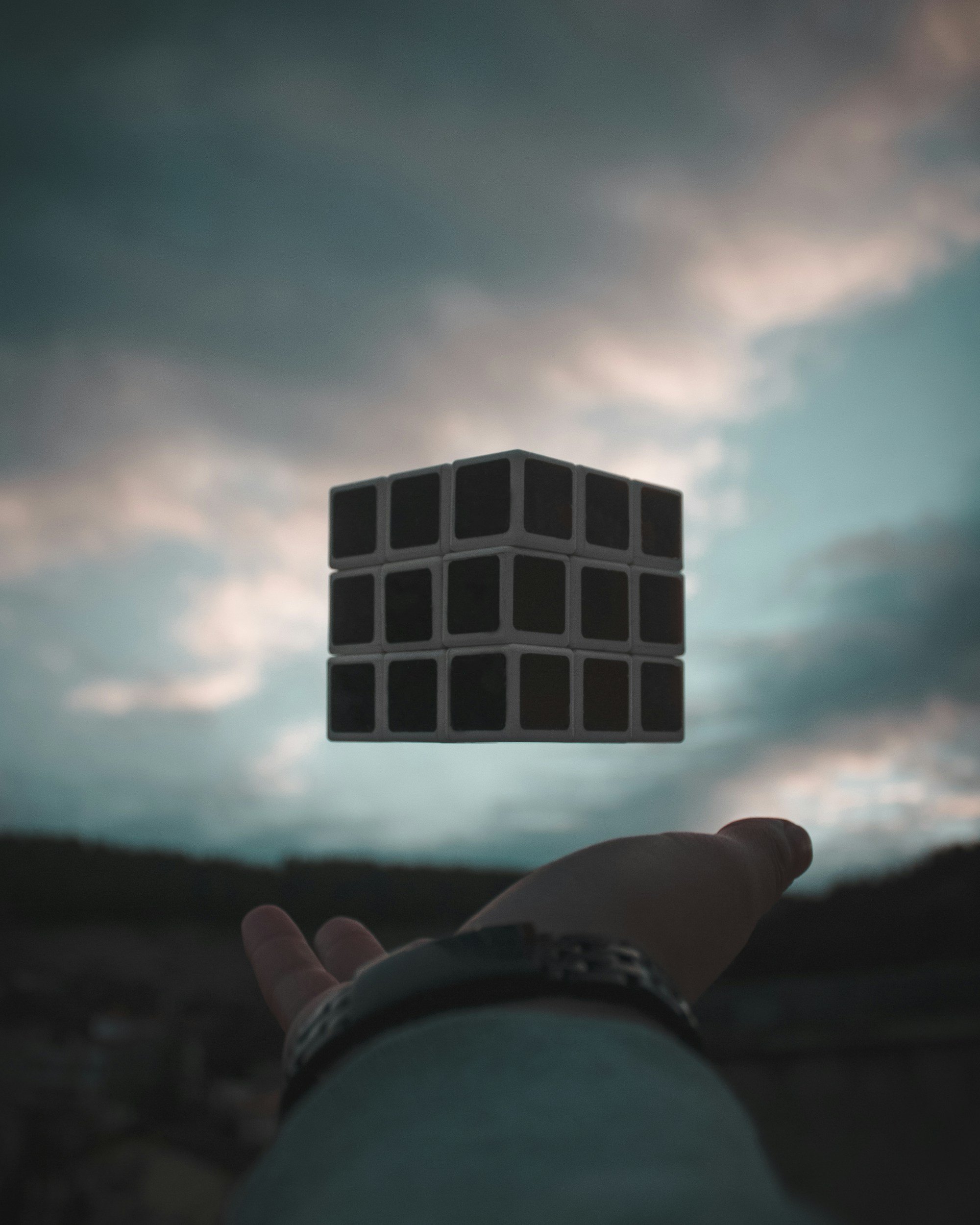 A hand extends toward the sky, with a floating, three-dimensional black cube with white borders in the background, set against a cloudy evening sky.