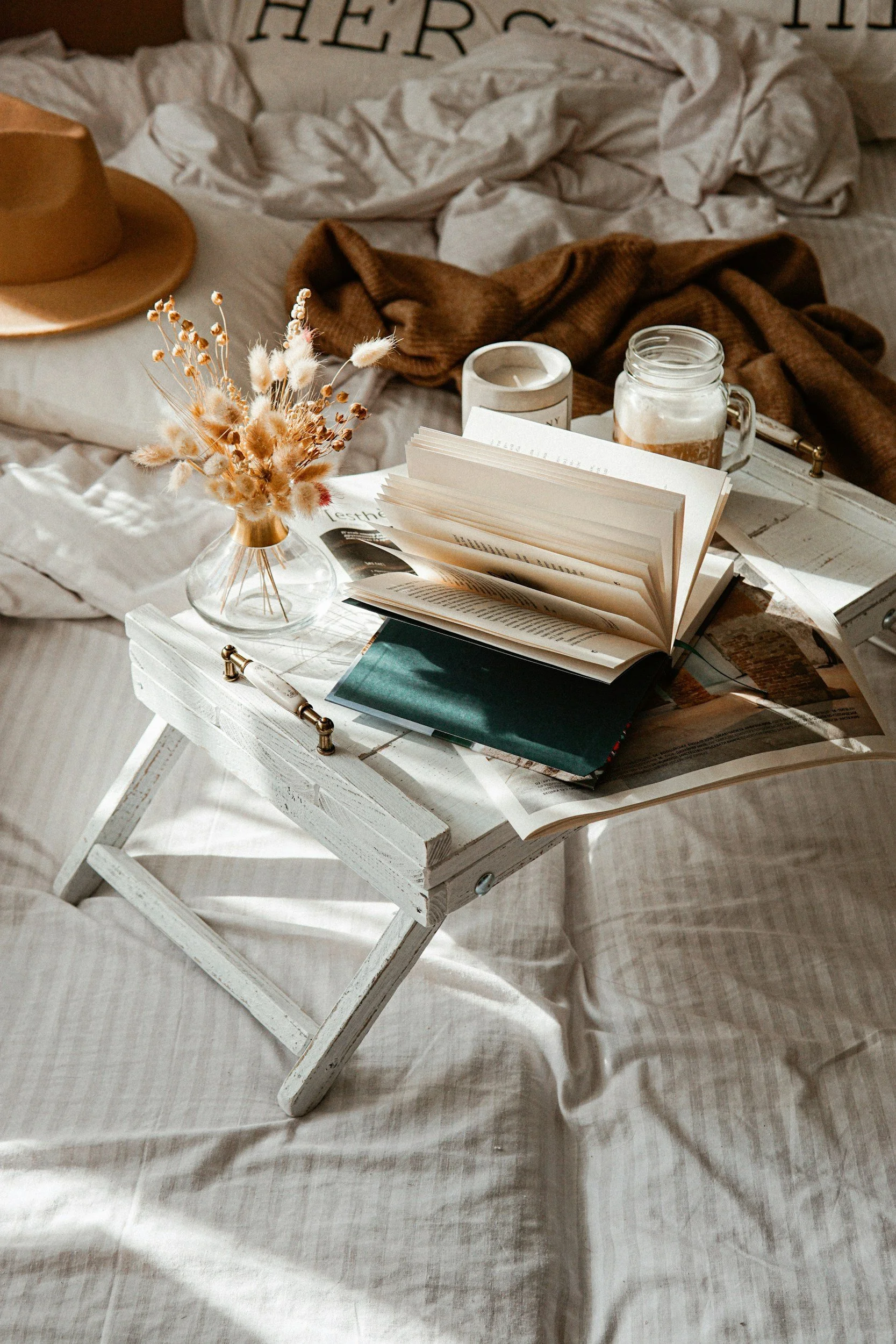 A small white wooden tray on a bed holding an open book, a closed book, and a magazine. There is a glass vase with dried flowers, a candle, a jar with a drink, and a pen nearby. The bed has beige and brown blankets, pillows, and a wide-brimmed hat.