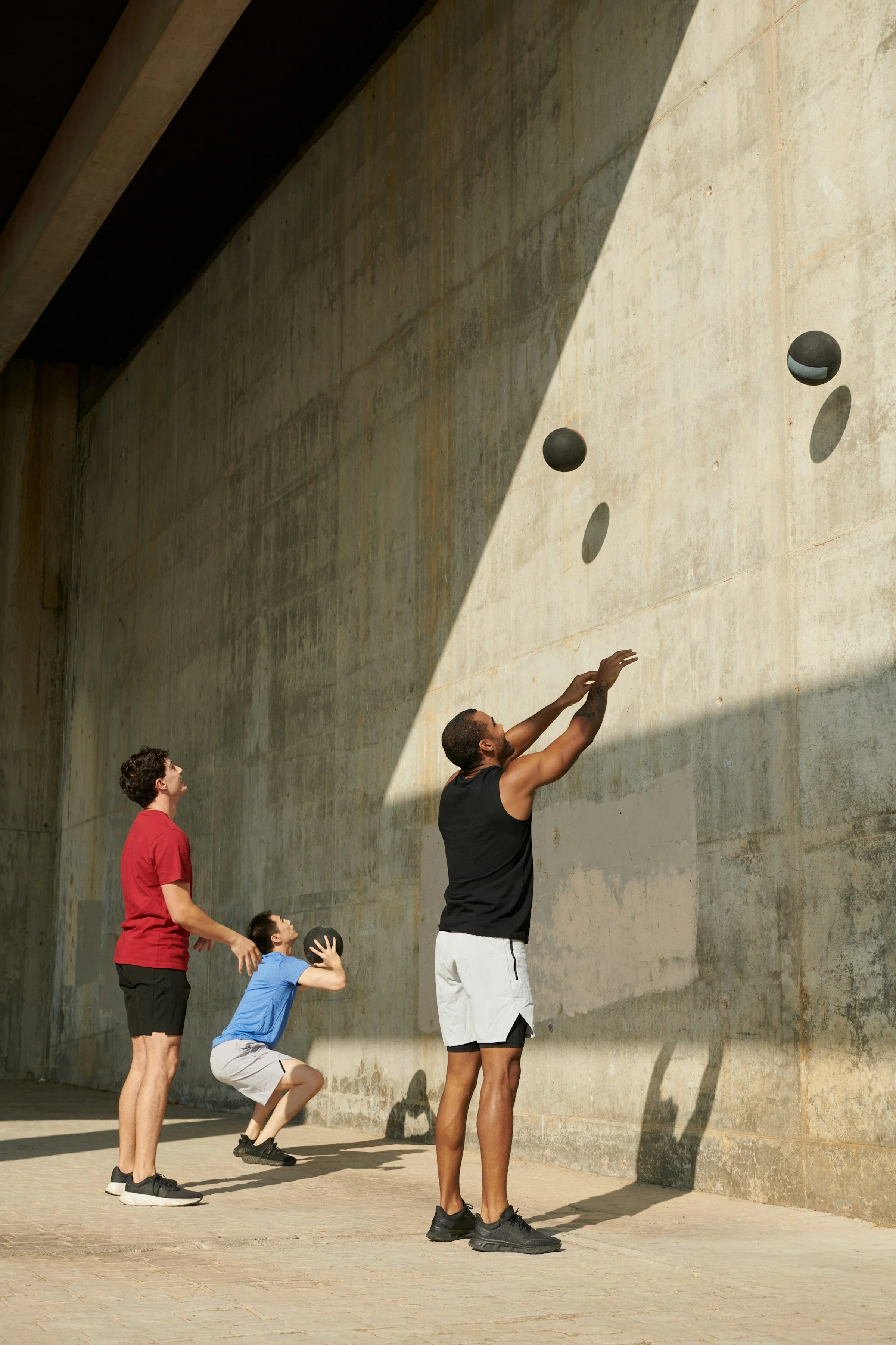 Three people playing basketball against a concrete wall with three black and white basketballs mounted on it. One person is shooting, while the other two are watching.