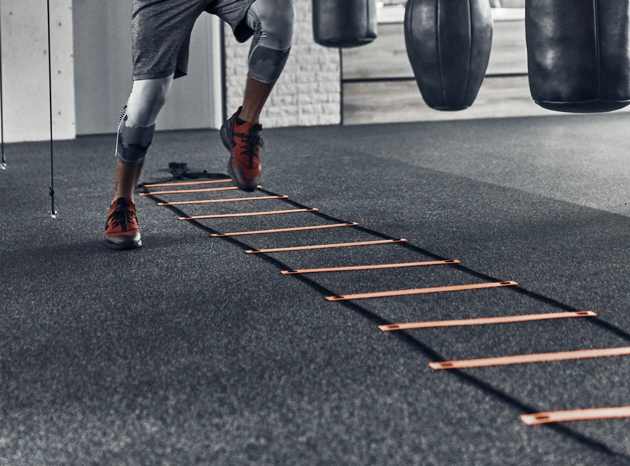 A person in athletic clothing and red running shoes is performing agility ladder drills on a rubber gym floor near punching bags.