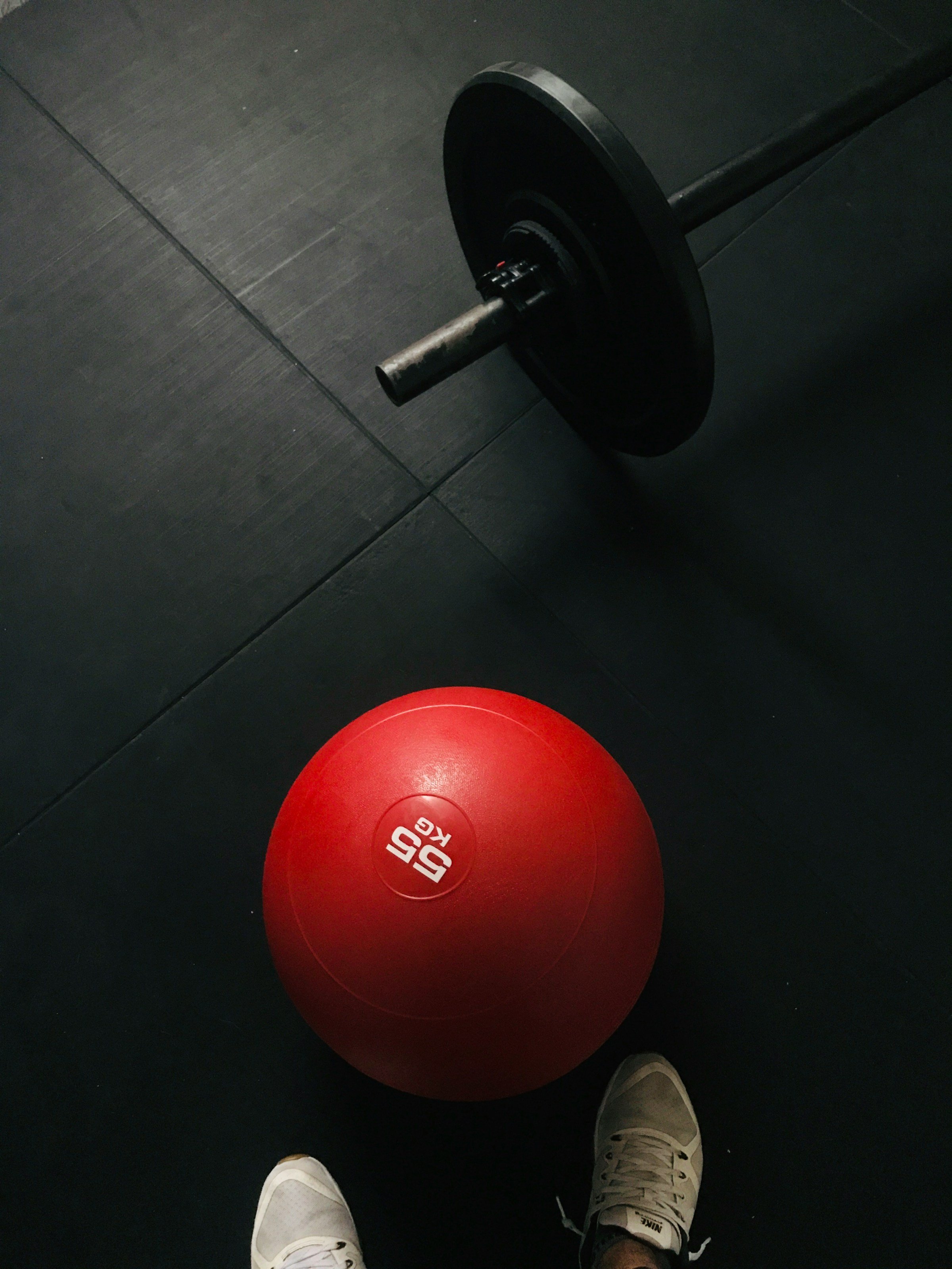 A black weightlifting barbell on the floor with a black weight plate attached, and a red exercise ball labeled '55 KG' on a gym floor, with person's white sneakers visible at the bottom.