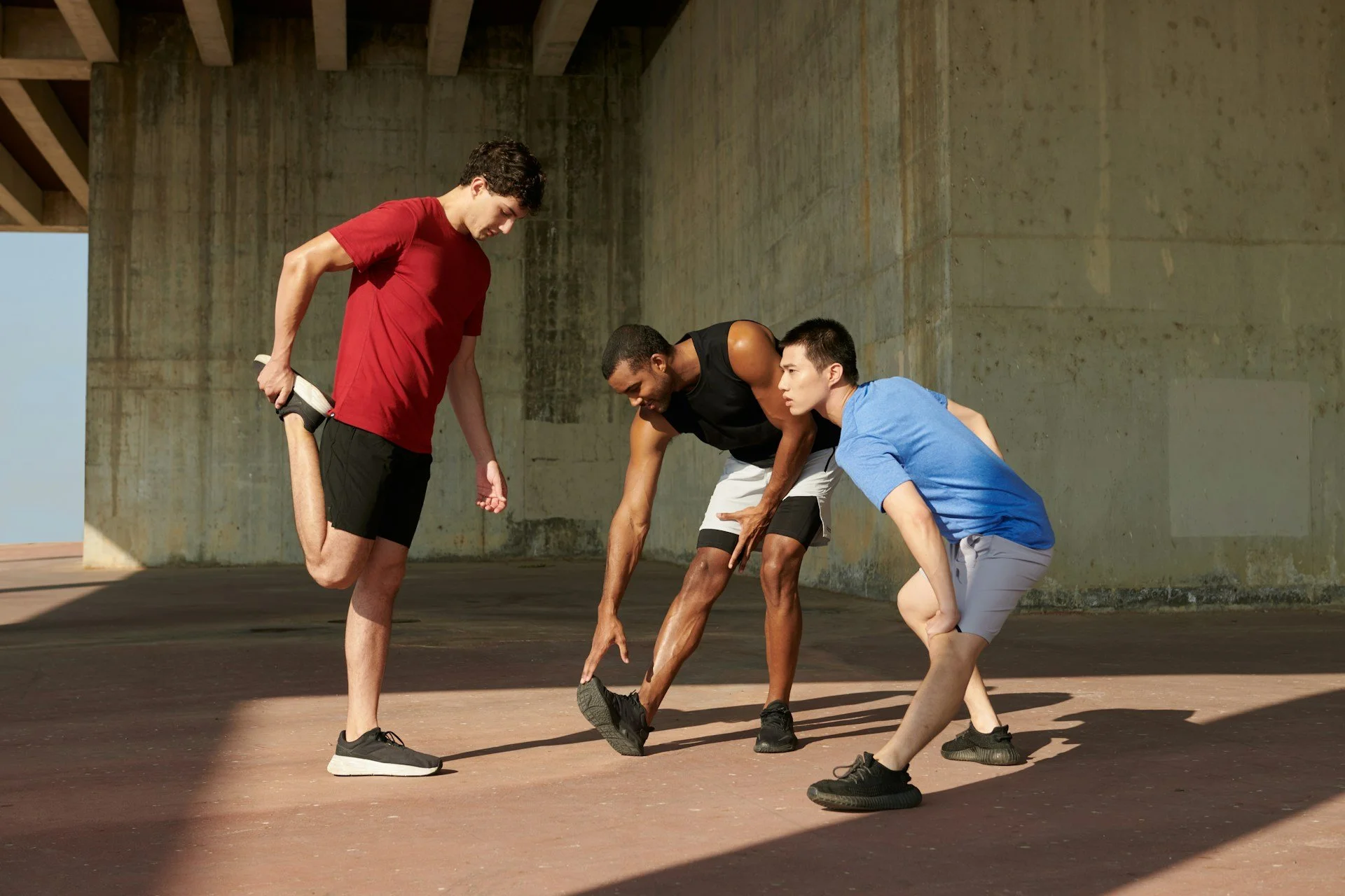 Three young men stretching outdoors under a concrete structure, with two men bending forward and one lifting his foot to his hand.