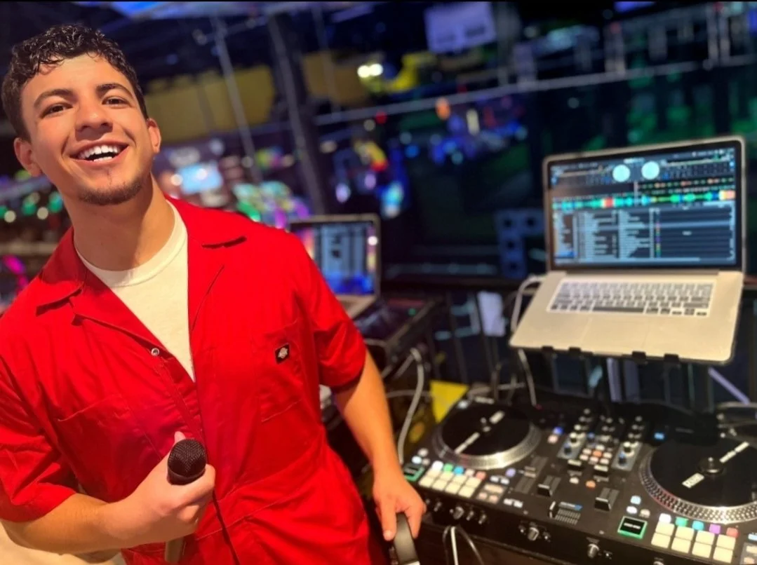 A young man smiling and holding a microphone at a DJ setup with laptop and DJ equipment, in a lively event space with colorful lights.