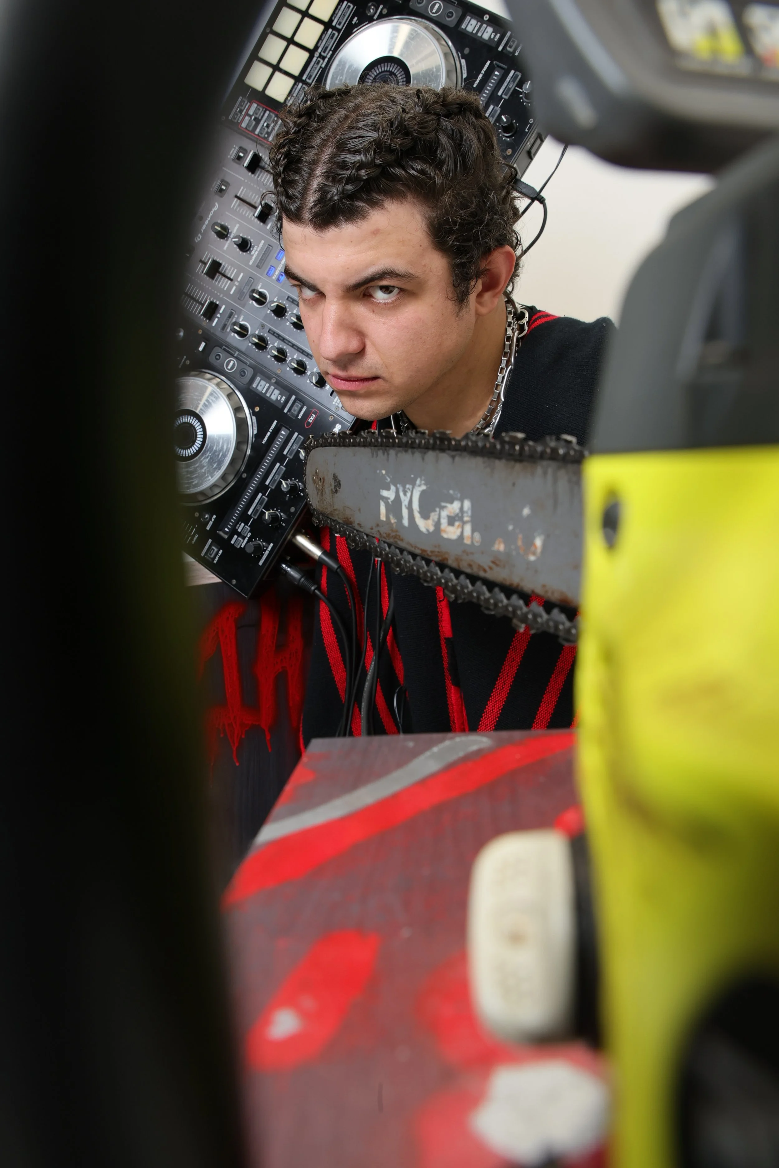 A person with dark curly hair and a silver chain wearing a black shirt with red accents, intently looking at the camera. He is surrounded by DJ equipment and what appears to be a chainsaw, all set in an indoor environment.