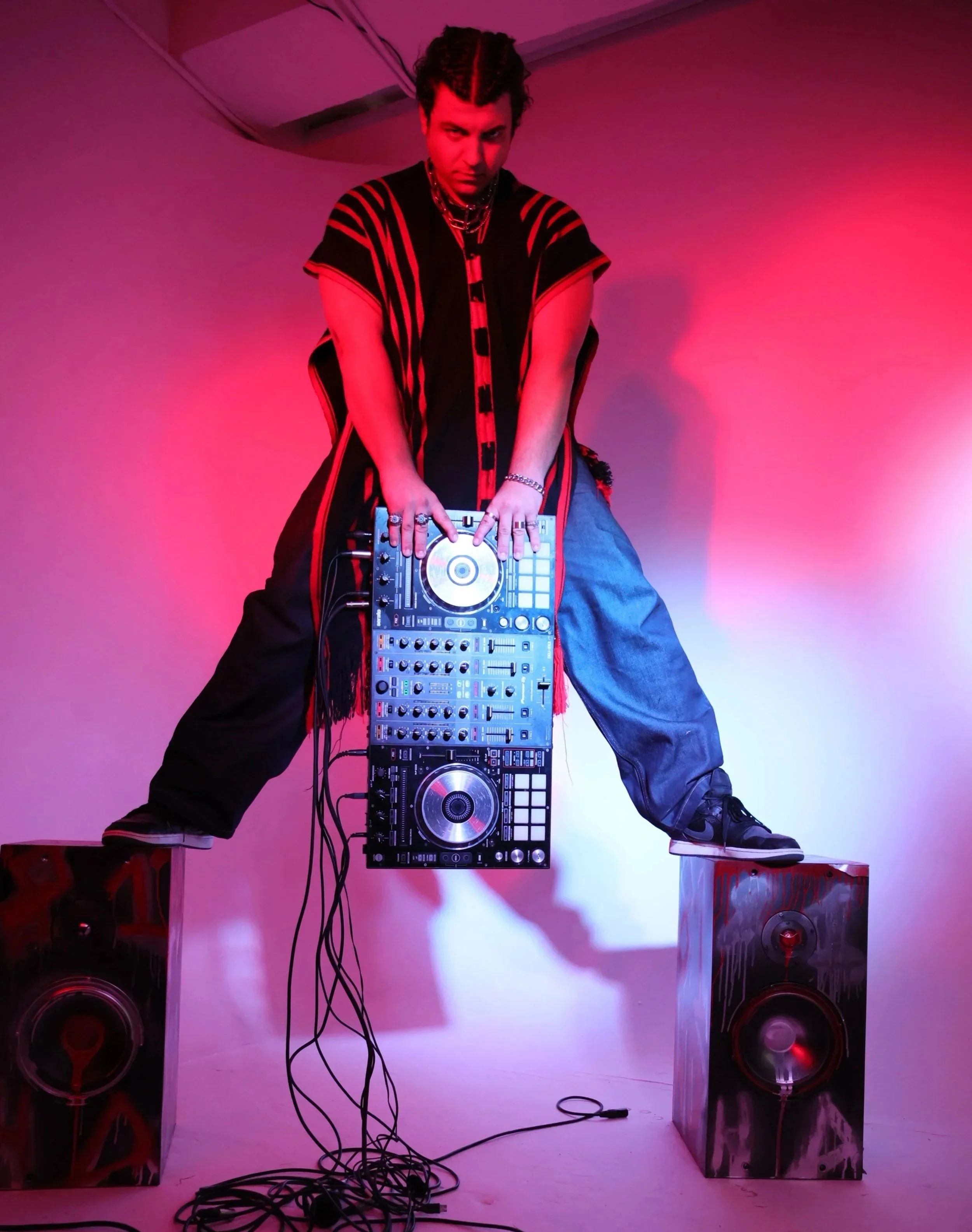 DJ standing on two speakers in a pink-lit room, leaning over a DJ controller, wearing baggy pants, a patterned vest, and jewelry.