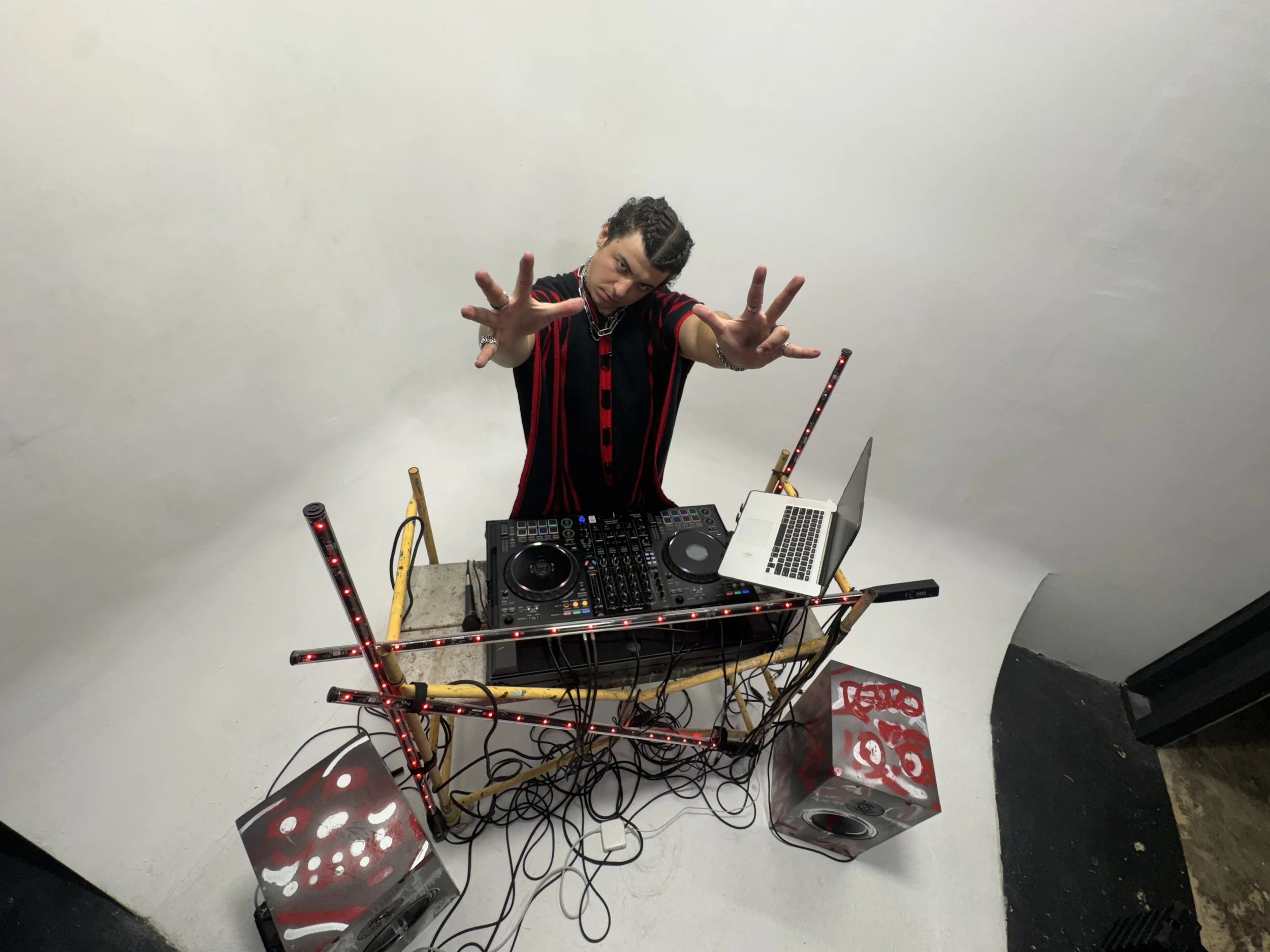 DJ standing behind a table with a DJ controller, laptop, and colorful LED lights, surrounded by speakers, in a studio with plain white walls.