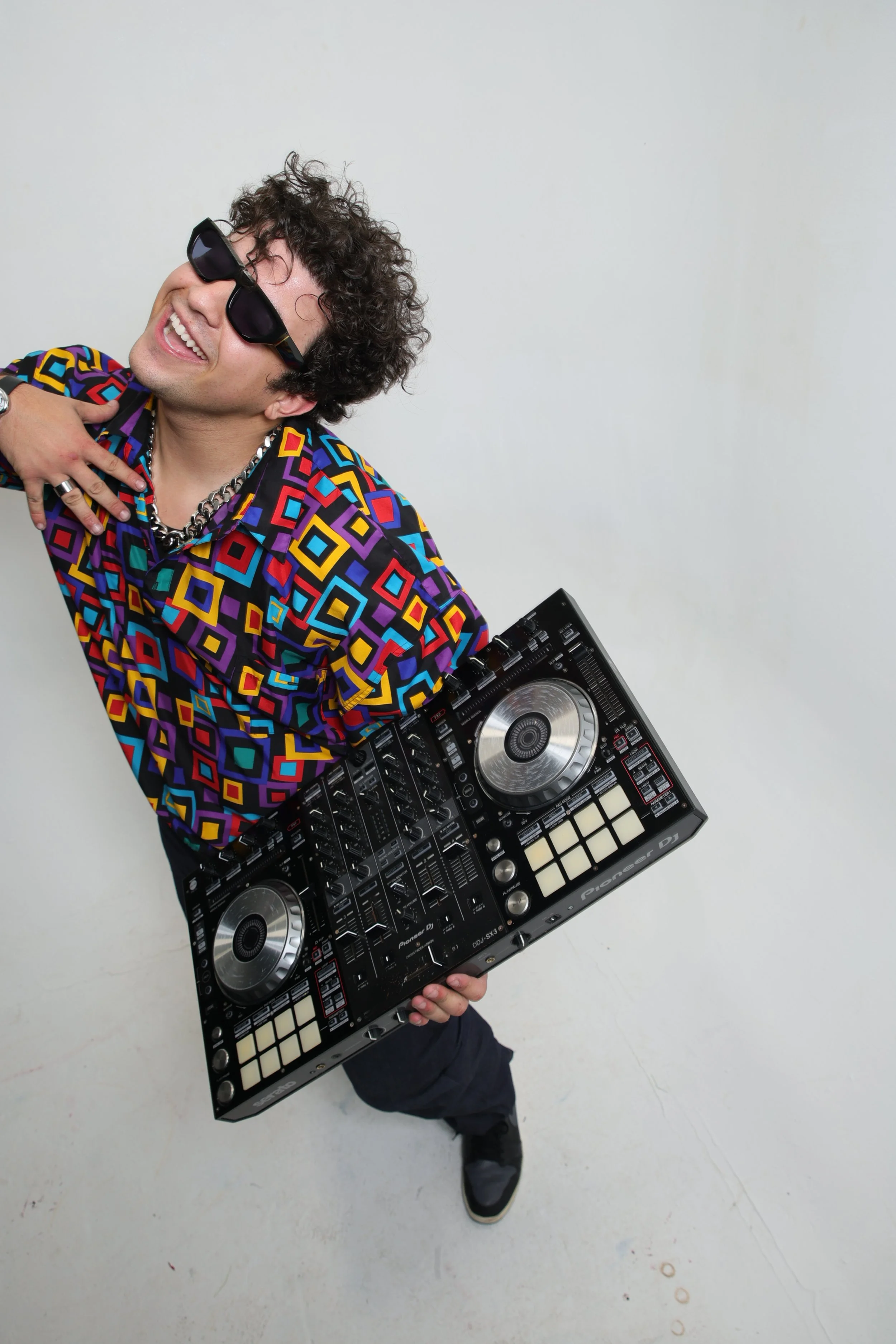 A young man with curly hair, wearing sunglasses, a colorful patterned shirt, and jewelry, holding a DJ controller and smiling while posing against a plain white background.