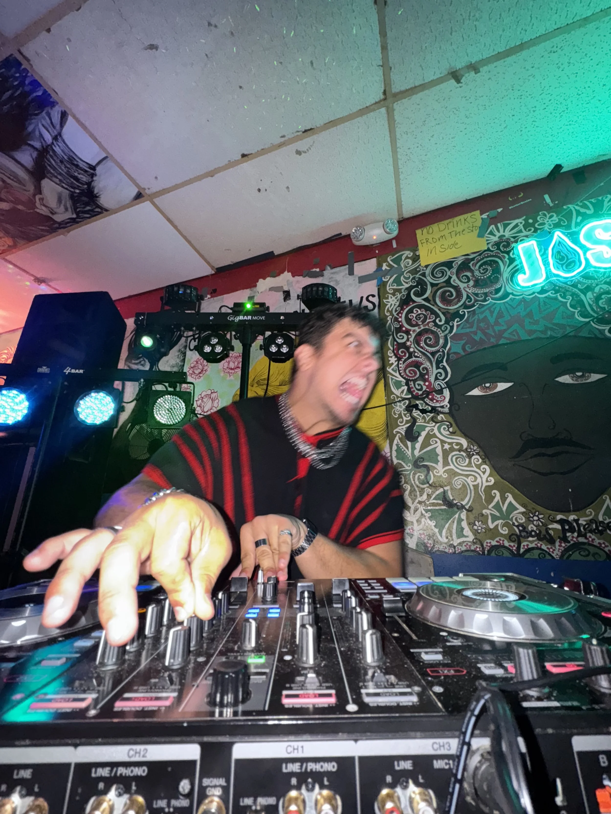A DJ in a black and red striped shirt performs at a nightclub, using a mixing console with colorful lights and speakers in the background. A colorful mural of a woman's face is visible on the wall, with a small yellow sign that reads 'NO DRINKS FROM 