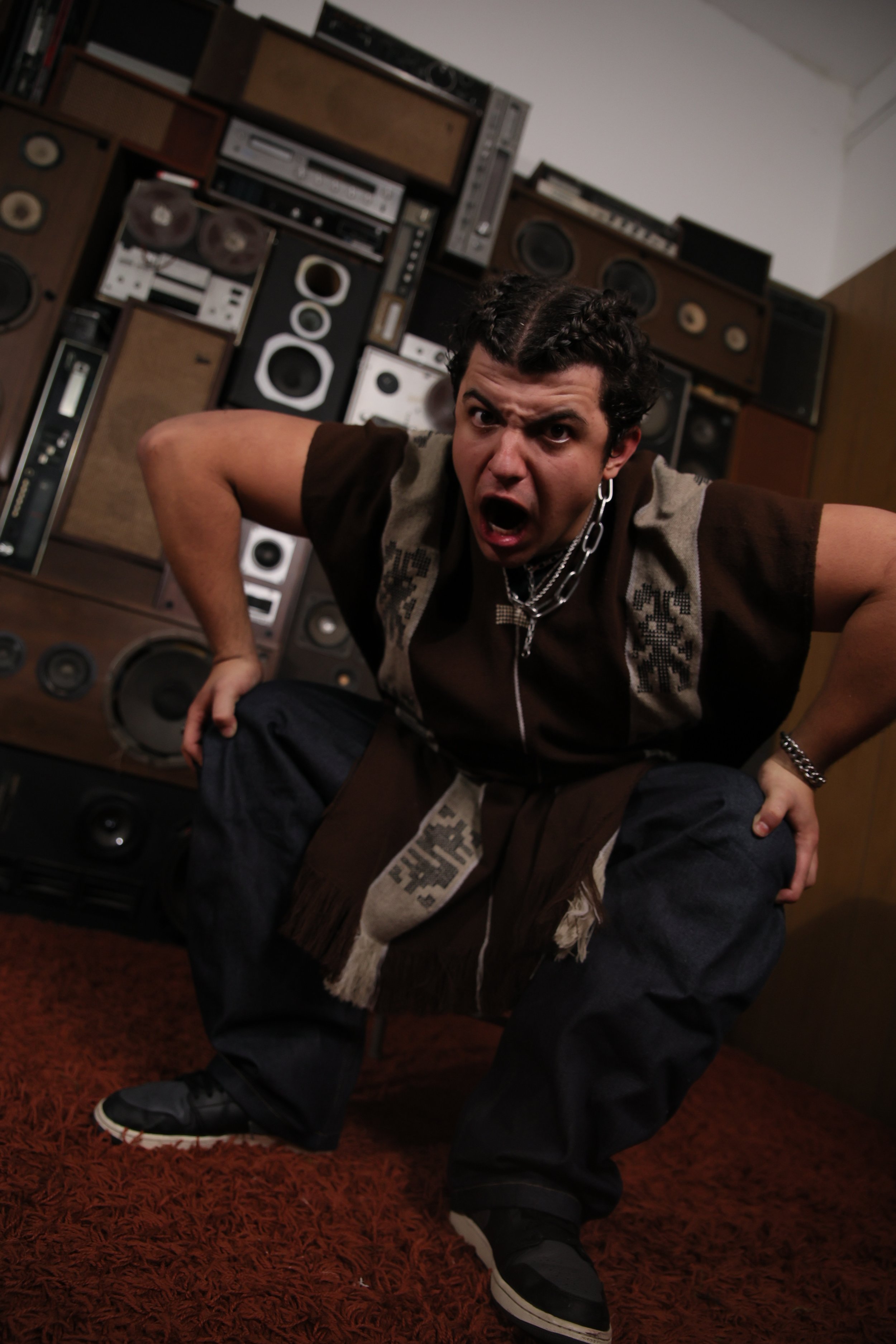 A man with dark, curly hair crouching in front of a large wall of vintage stereo equipment, wearing a brown and beige knitted vest, dark pants, and black sneakers, with an intense expression and mouth open.