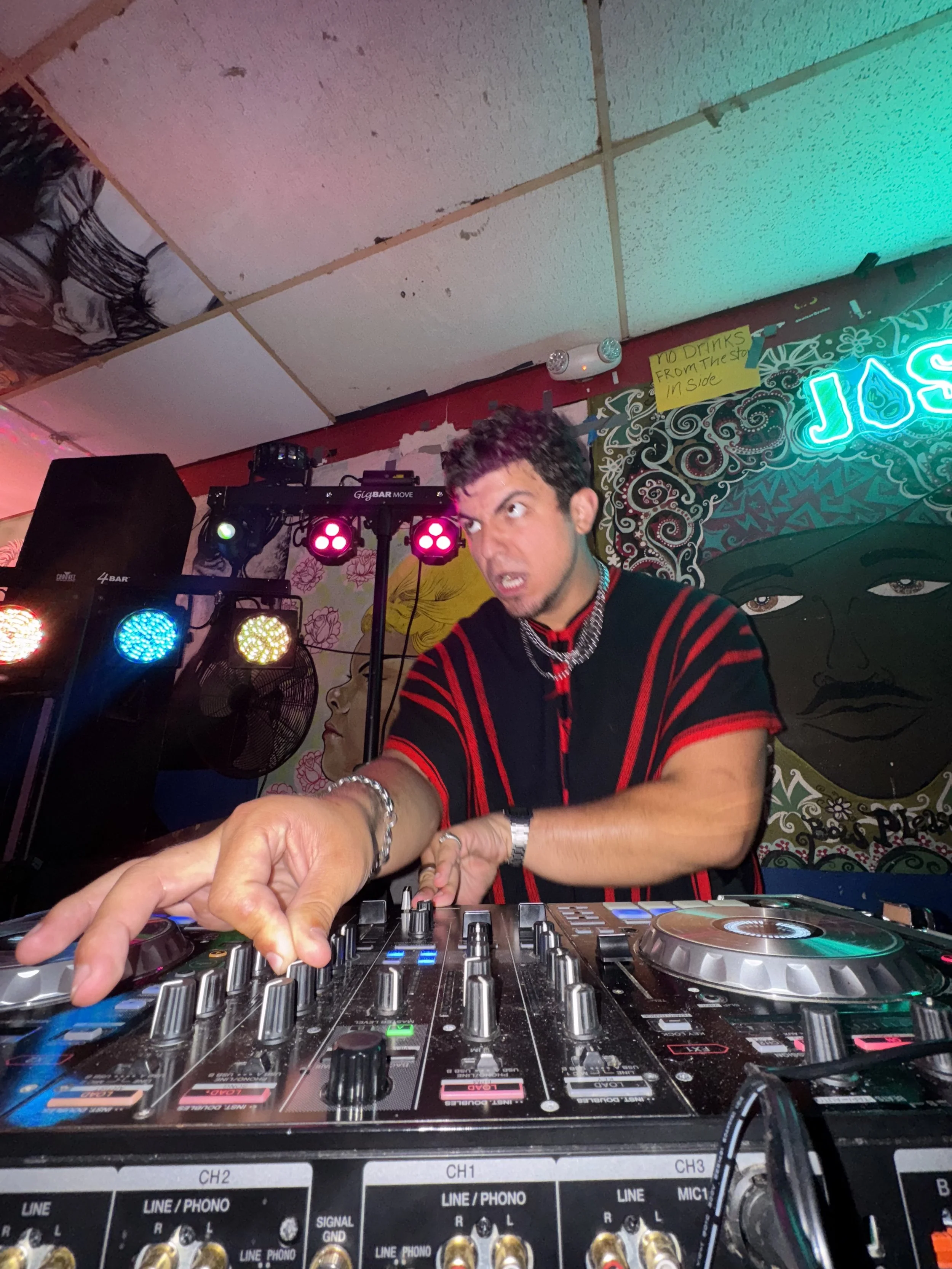 A DJ is performing behind a mixing console at a nightclub or bar, with colorful lights and a mural featuring a woman's face and artistic patterns in the background.