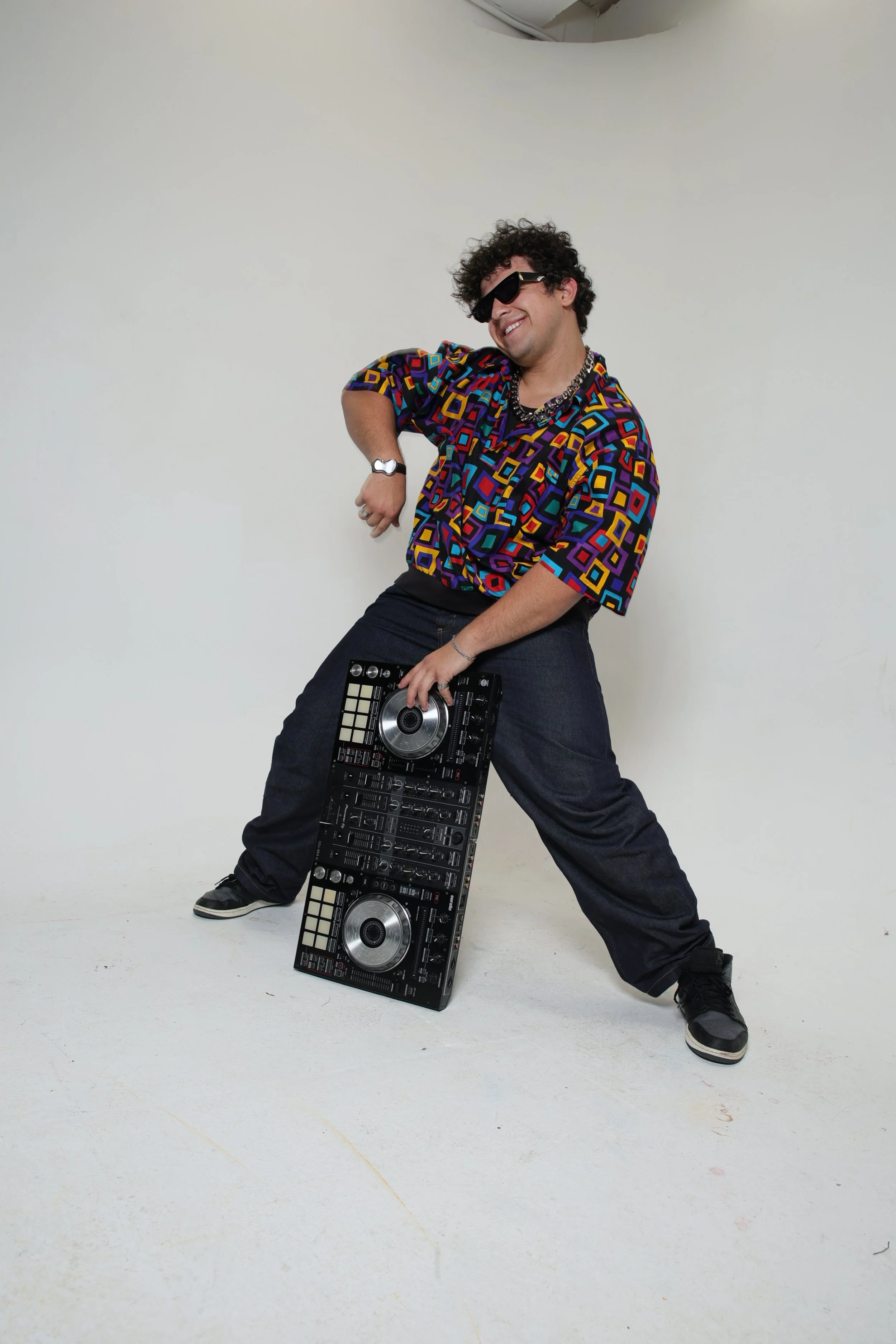 A man dressed in colorful geometric patterned shirt, black pants, and sneakers, is posing with a DJ controller on the floor, smiling and wearing sunglasses in a studio with a plain light background.