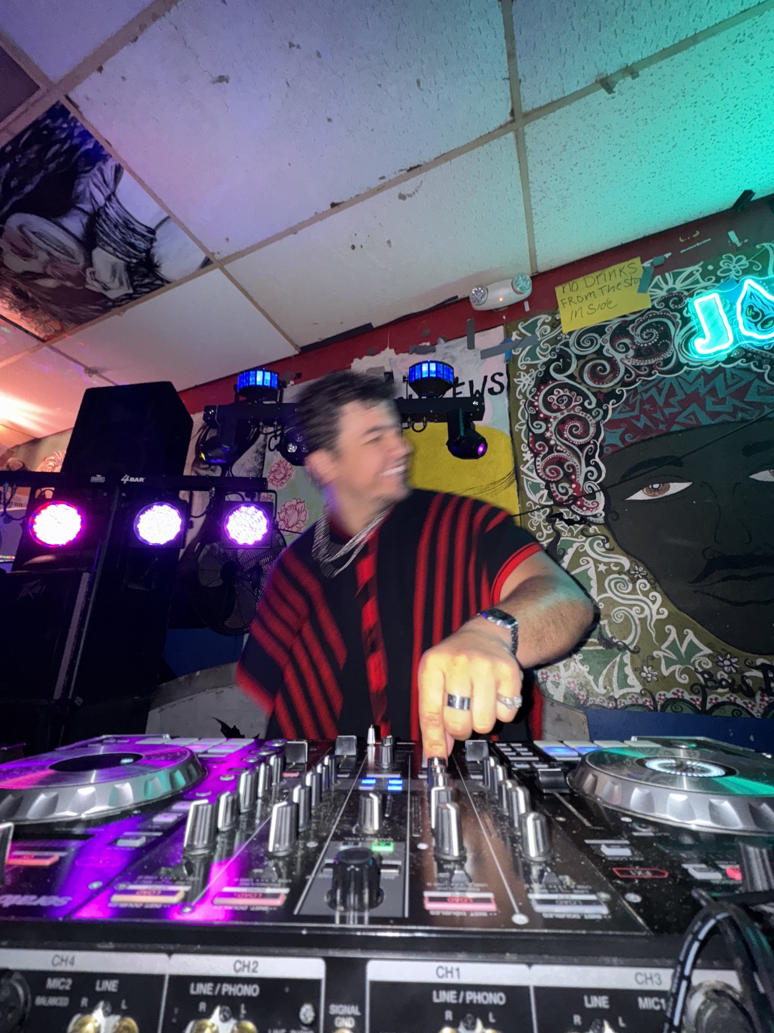 A DJ wearing a red and black striped shirt, smiling and pointing at a DJ mixer with colorful lights, in a vibrant, decorated venue with artwork and signage on the wall.