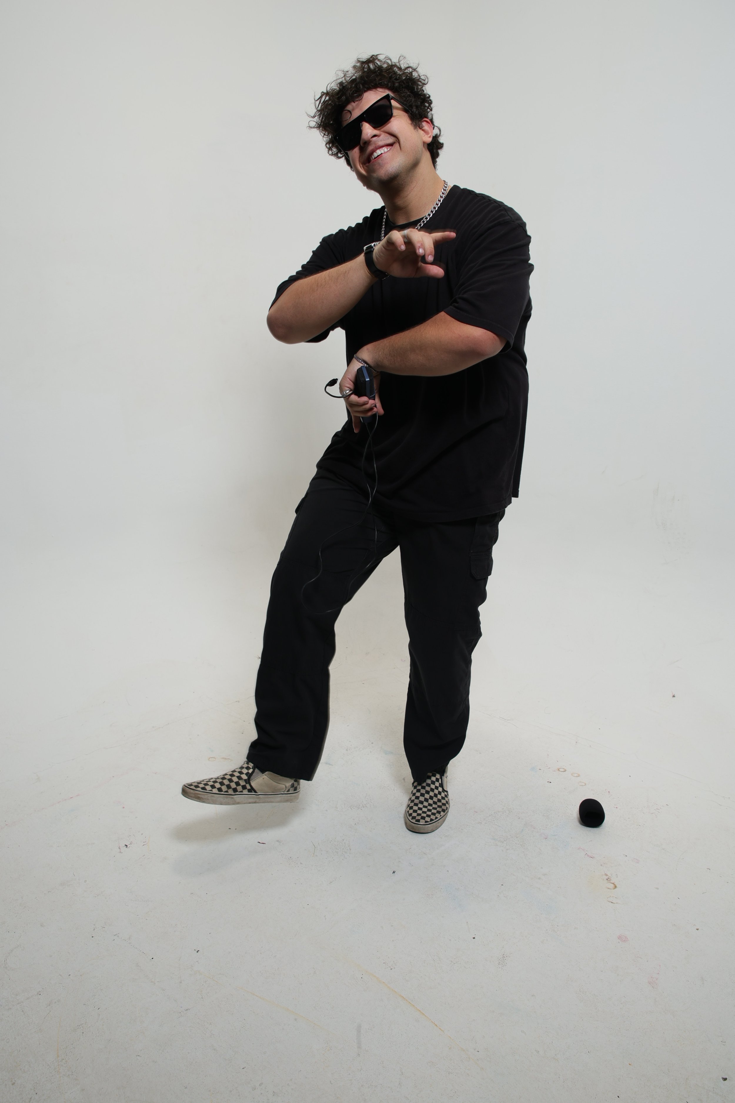 A young man with curly hair wearing sunglasses, a black t-shirt, black pants, checkered slip-on shoes, and accessories, smiling and holding a walkie-talkie, standing against a plain white background.