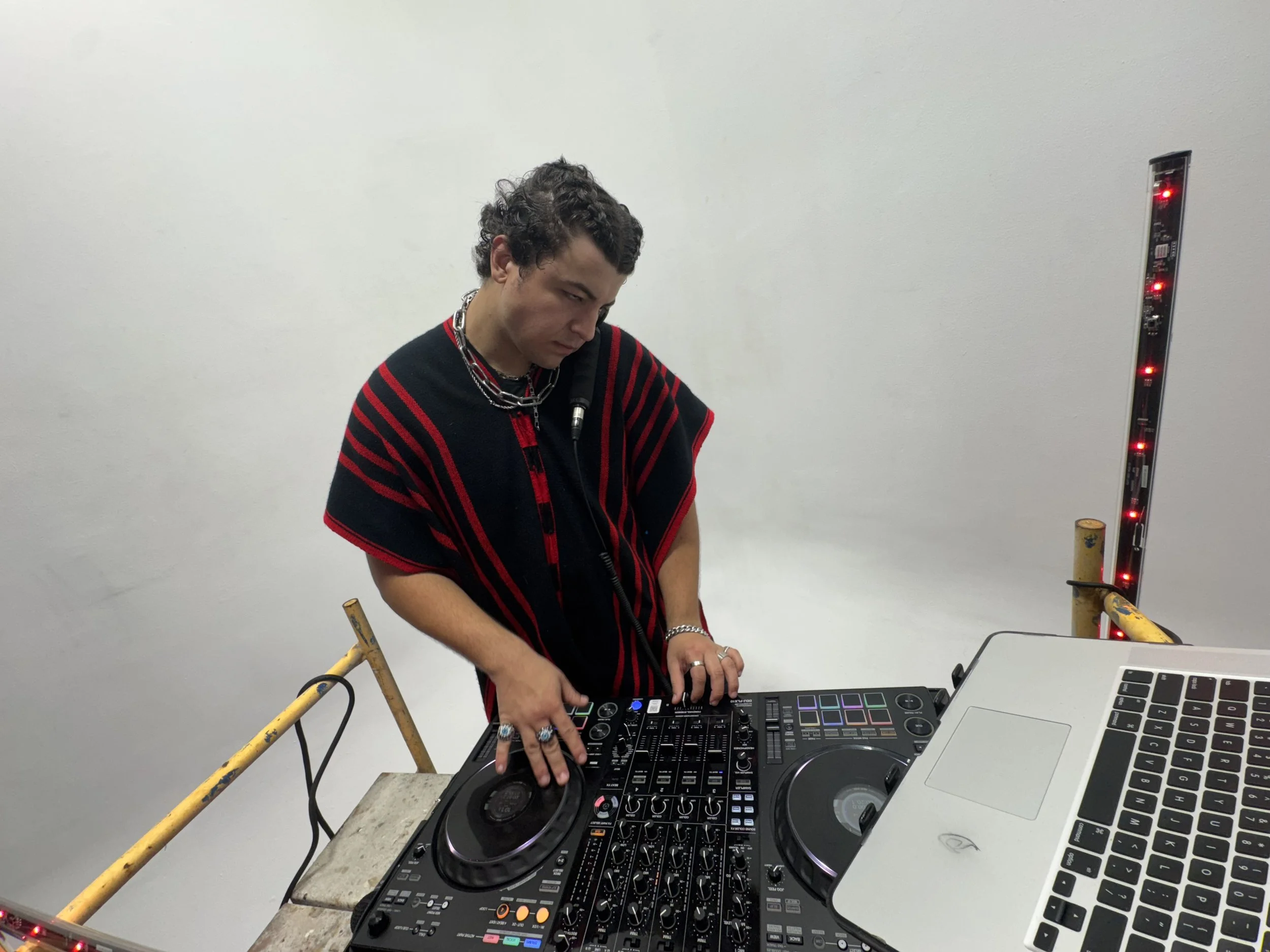 A person with curly hair wearing a black and red striped top, standing behind a DJ controller and a laptop, in a minimalistic setting with white walls and some standing lighting with red lights.