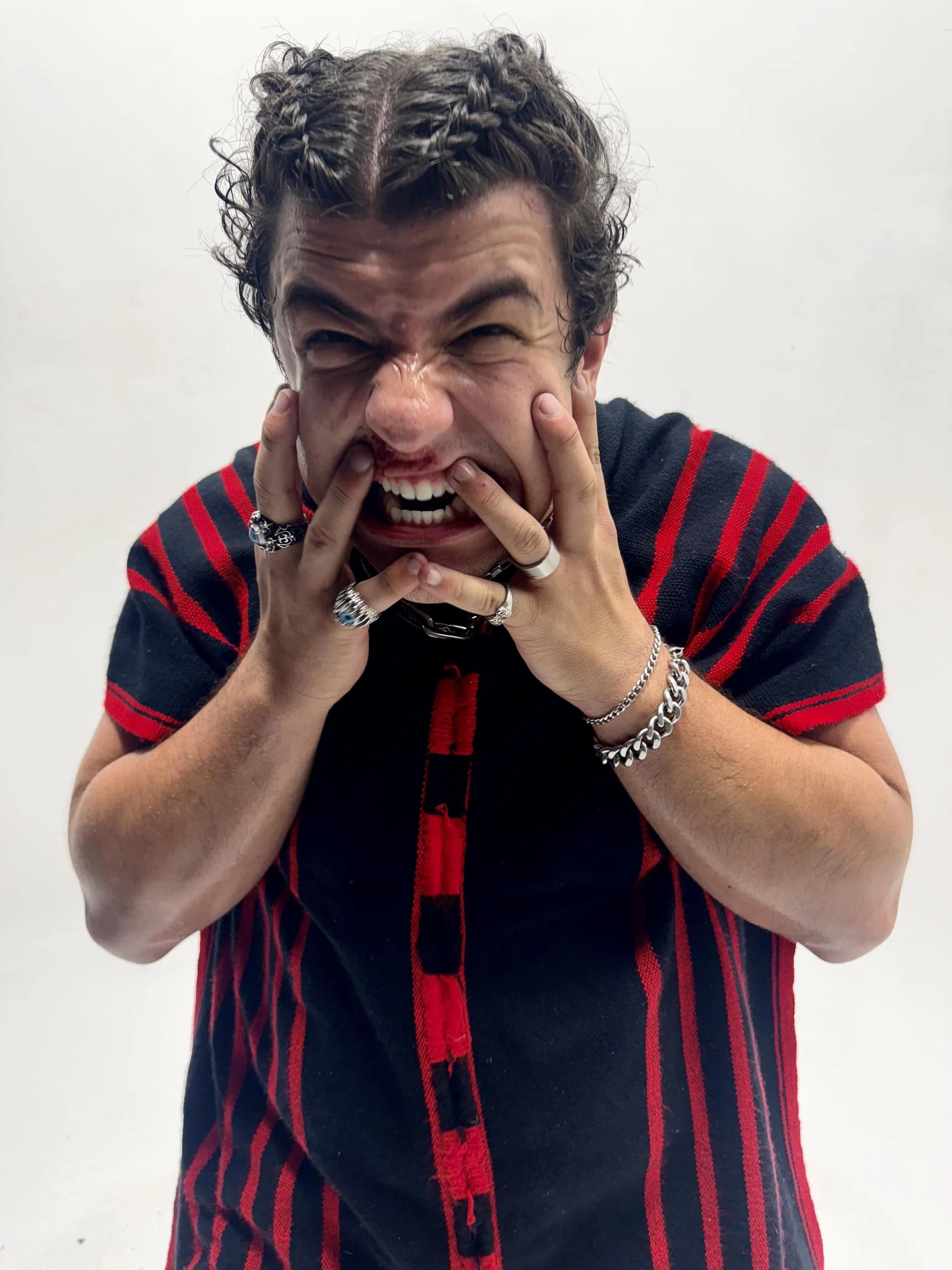A man with an intense, angry expression, pulling his face with his hands, showing clenched teeth, and wearing multiple rings and chains, against a plain white background.