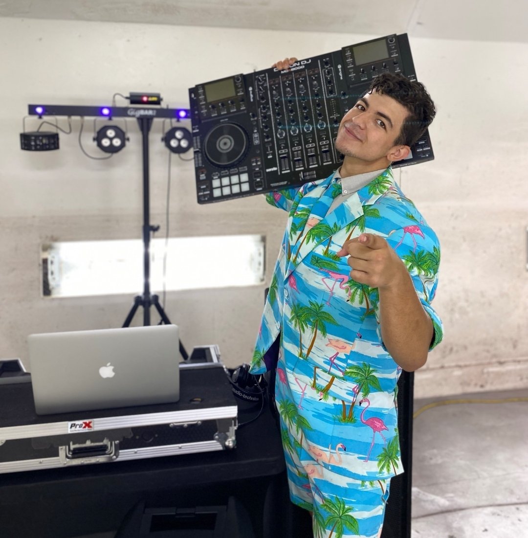 A man in a colorful Hawaiian shirt holds a DJ controller over his shoulder, smiling and pointing at the camera in a room with DJ equipment and a lighting setup.