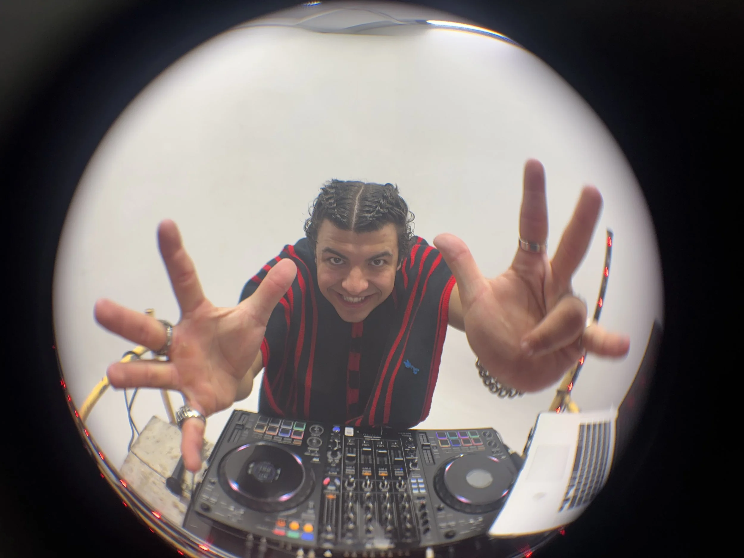 A DJ with dark, curly hair smiling and reaching towards the camera, standing behind a DJ mixer and equipment, seen through a fisheye lens.