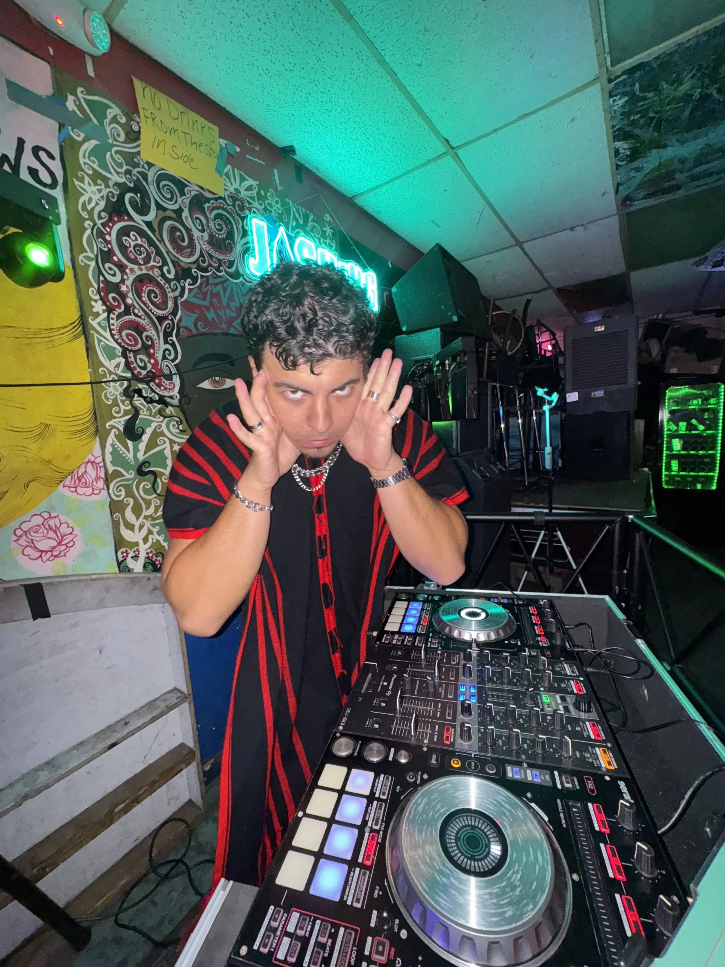 A DJ with curly hair and jewelry is posing behind a DJ setup with turntables and a mixer in a nightclub or bar. There are colorful lights and artwork on the wall behind him.