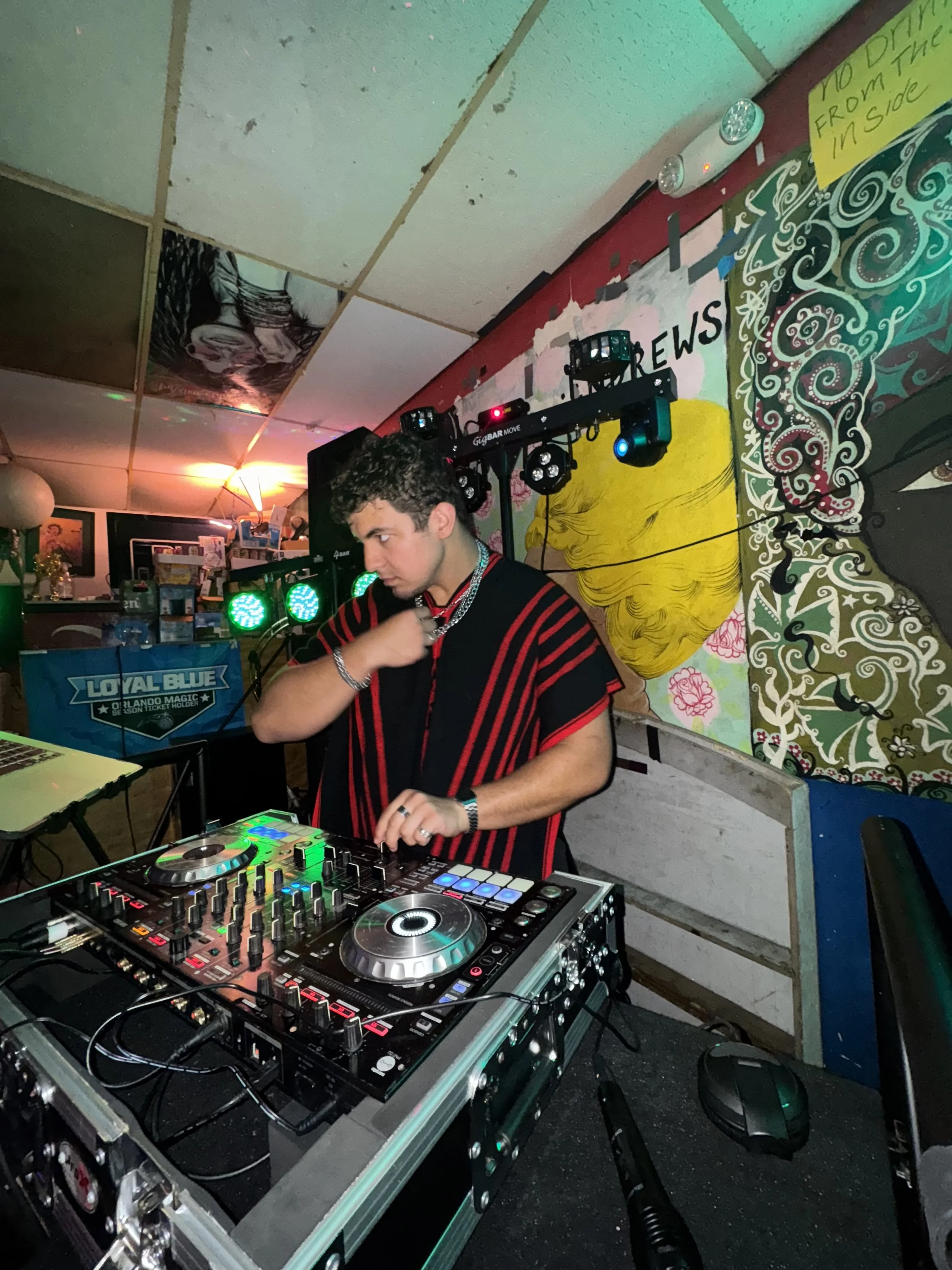 A DJ with short dark curly hair wearing a black and red striped shirt is performing at a party, mixing music on a DJ controller. The background features colorful graffiti-style artwork and a lighting setup with green and blue lights.