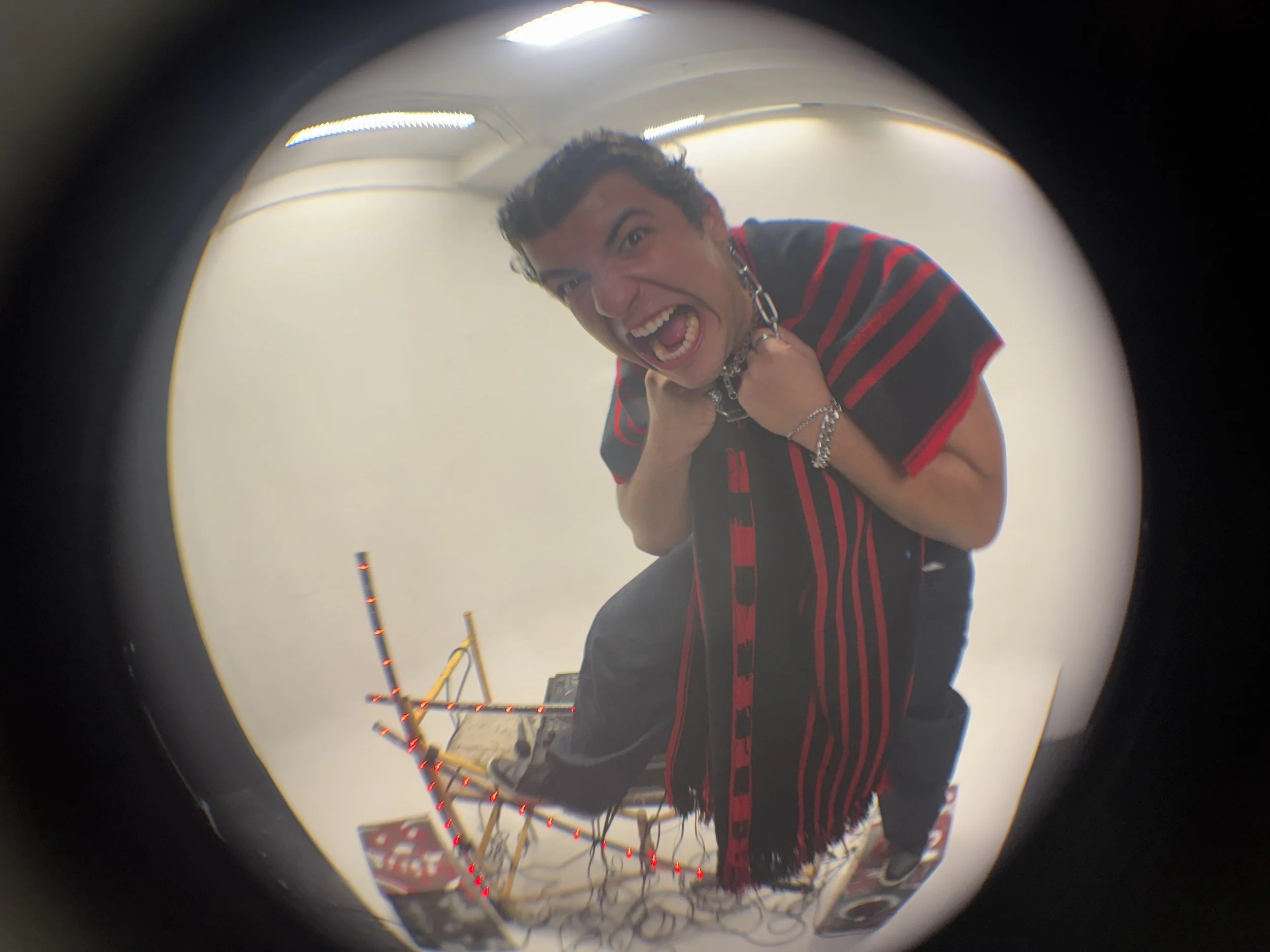 A young man with dark hair making an intense, fierce facial expression, shouting with mouth open wide, eyes wide open, and wearing a black shirt with red stripes, jewelry, and a scarf. He is kneeling and holding a chain around his neck, with a white 