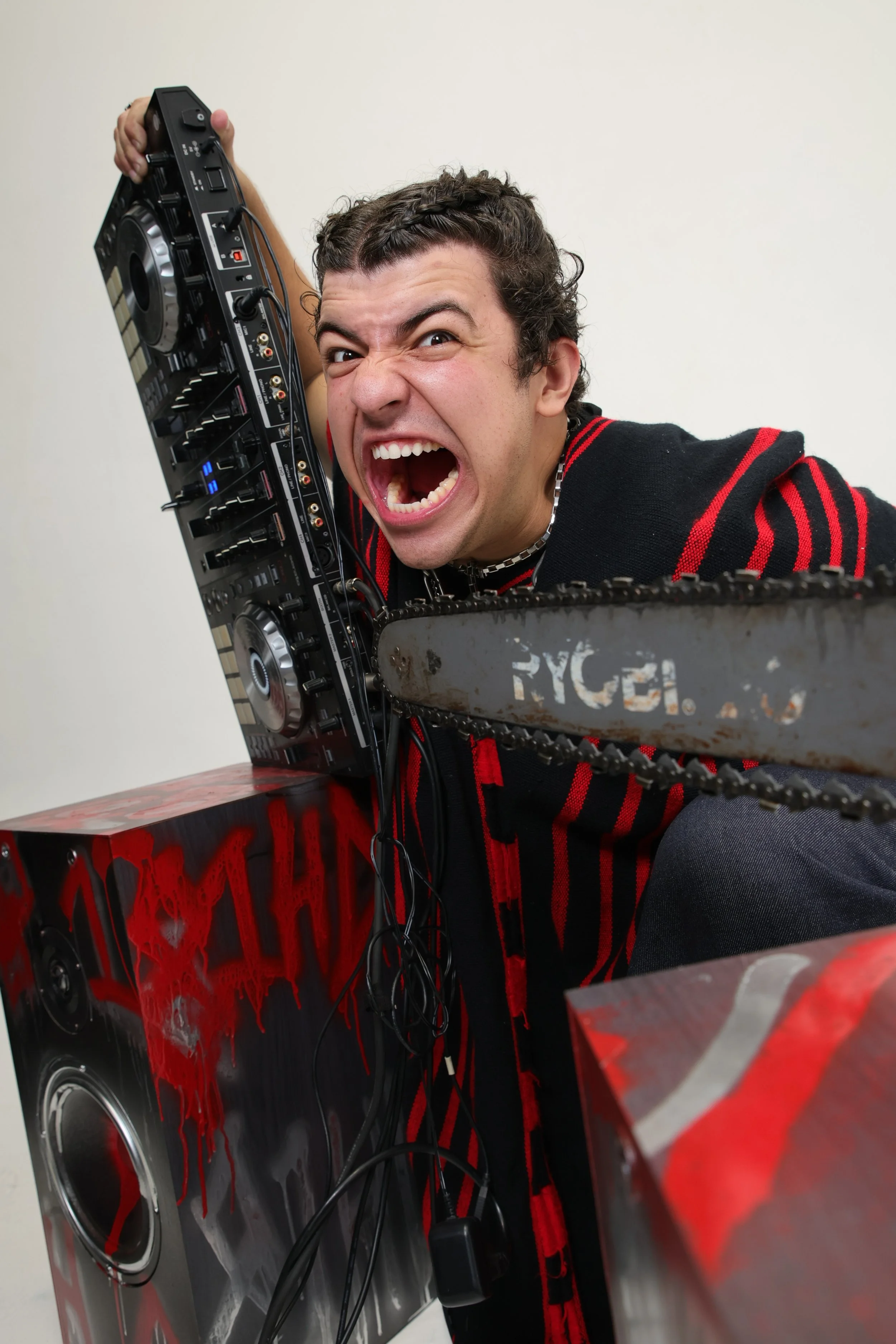 A man with curly hair and a beaded necklace making an angry or intense face, holding DJ equipment, with red and black painted speakers around him.