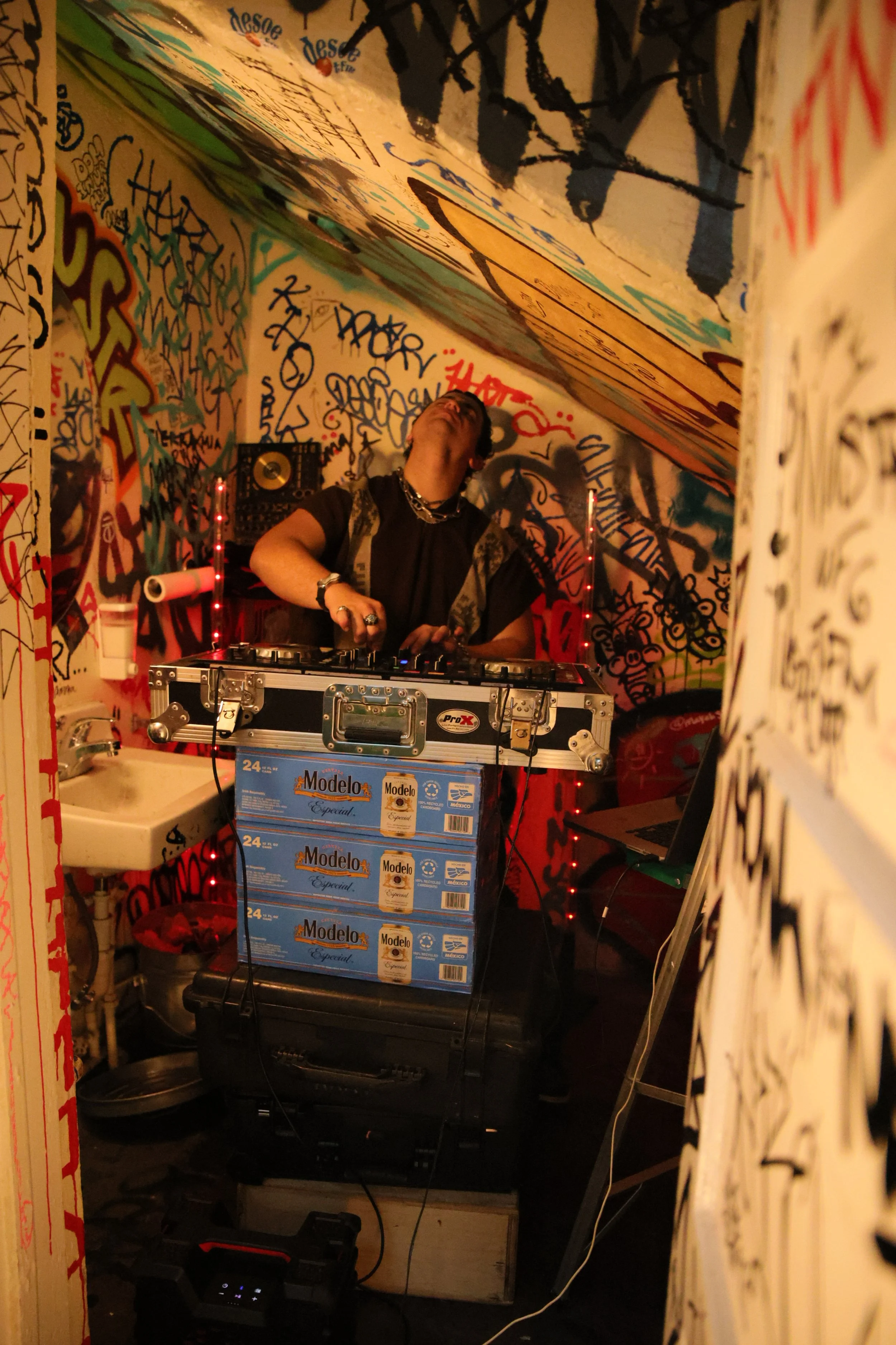 Person DJing in a graffiti-covered room with a turntable, boxes of Modelo beer, and red string lights.