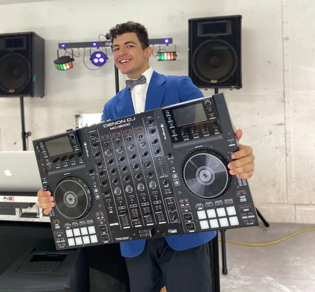 A young man dressed in a blue blazer and gray bow tie smiling while holding a large DJ controller. Behind him are speakers and colorful lighting equipment in a modern indoor setting.