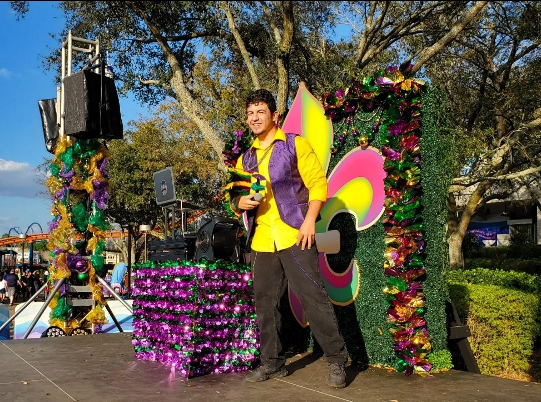A young man dressed in a yellow shirt and purple vest smiling on a decorated float with colorful butterfly wings, Mardi Gras beads, and a green festive backdrop, outdoors during daytime.