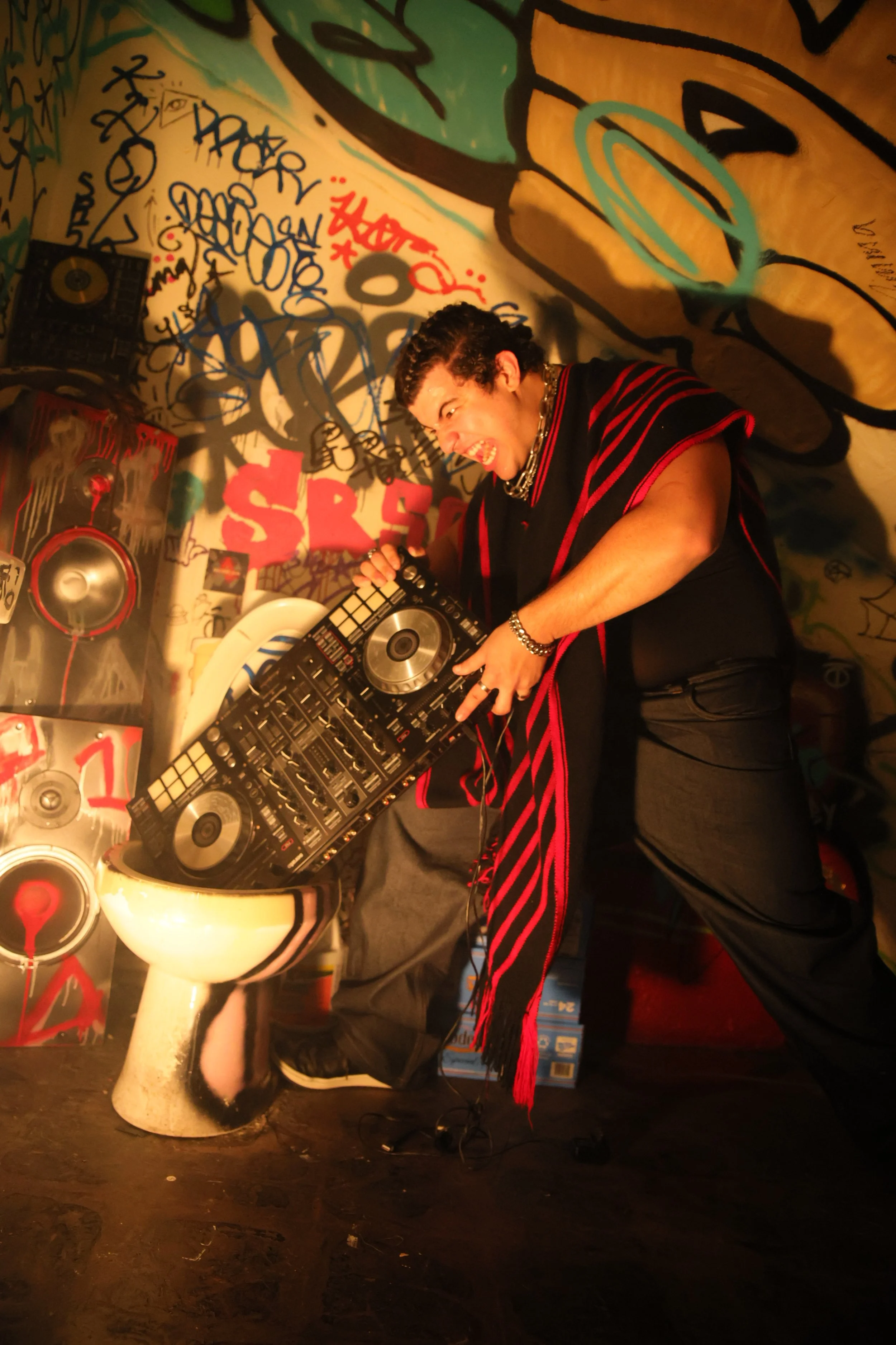A person with curly hair and dark clothing, wearing silver jewelry, is acting like they are performing as a DJ with a DJ controller, with a playful and intense expression. They are standing on a dark speckled floor in front of graffiti-covered walls 