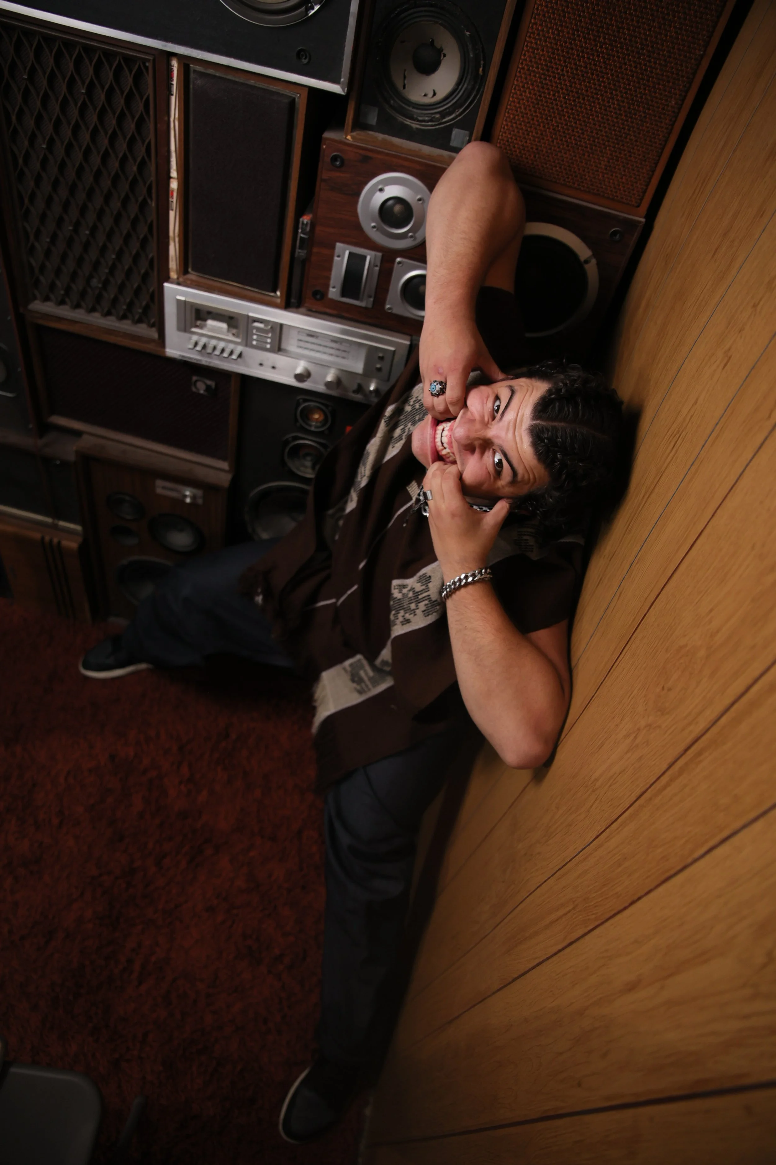 A man lying on a wooden floor, surrounded by large vintage speakers, smiling and looking up at the camera.