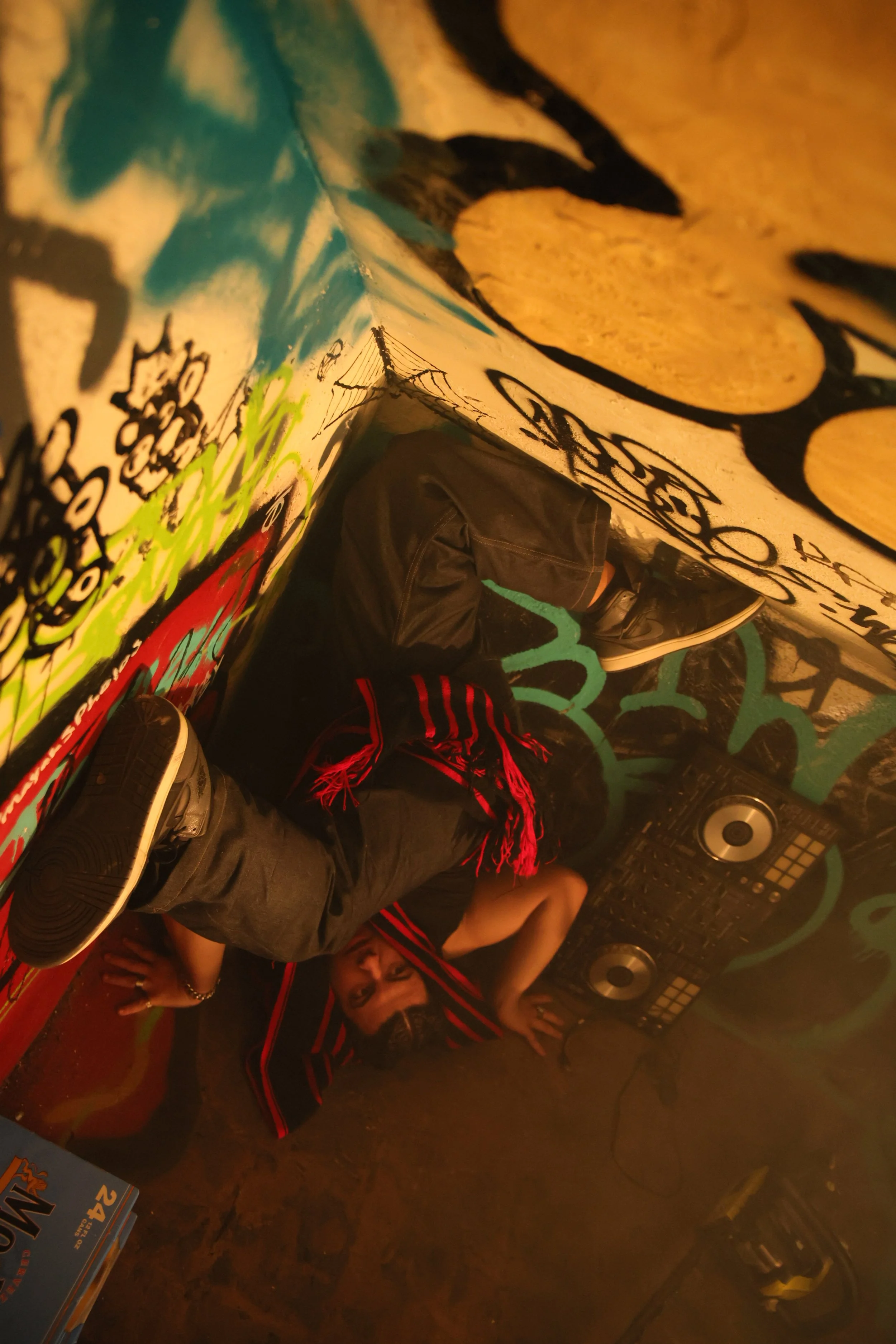 Person lying on the floor in a graffiti-covered corner, surrounded by DJ equipment, with a casual outfit and red and black scarf.