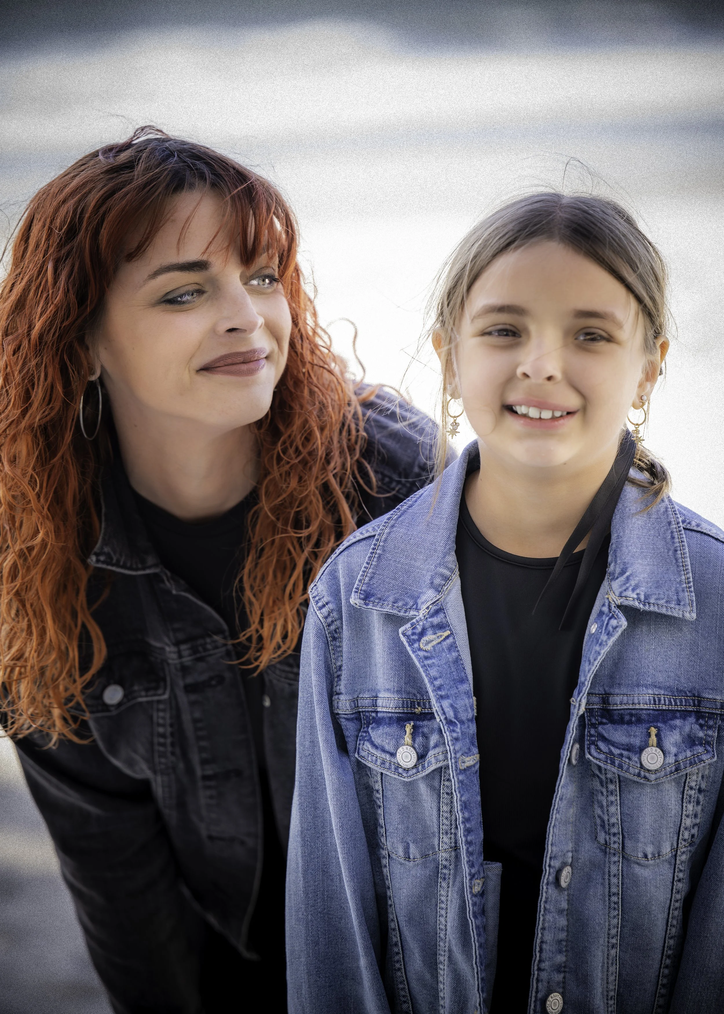 A woman with curly red hair and a young girl with brown hair in pigtails wearing denim jackets standing close together outdoors.