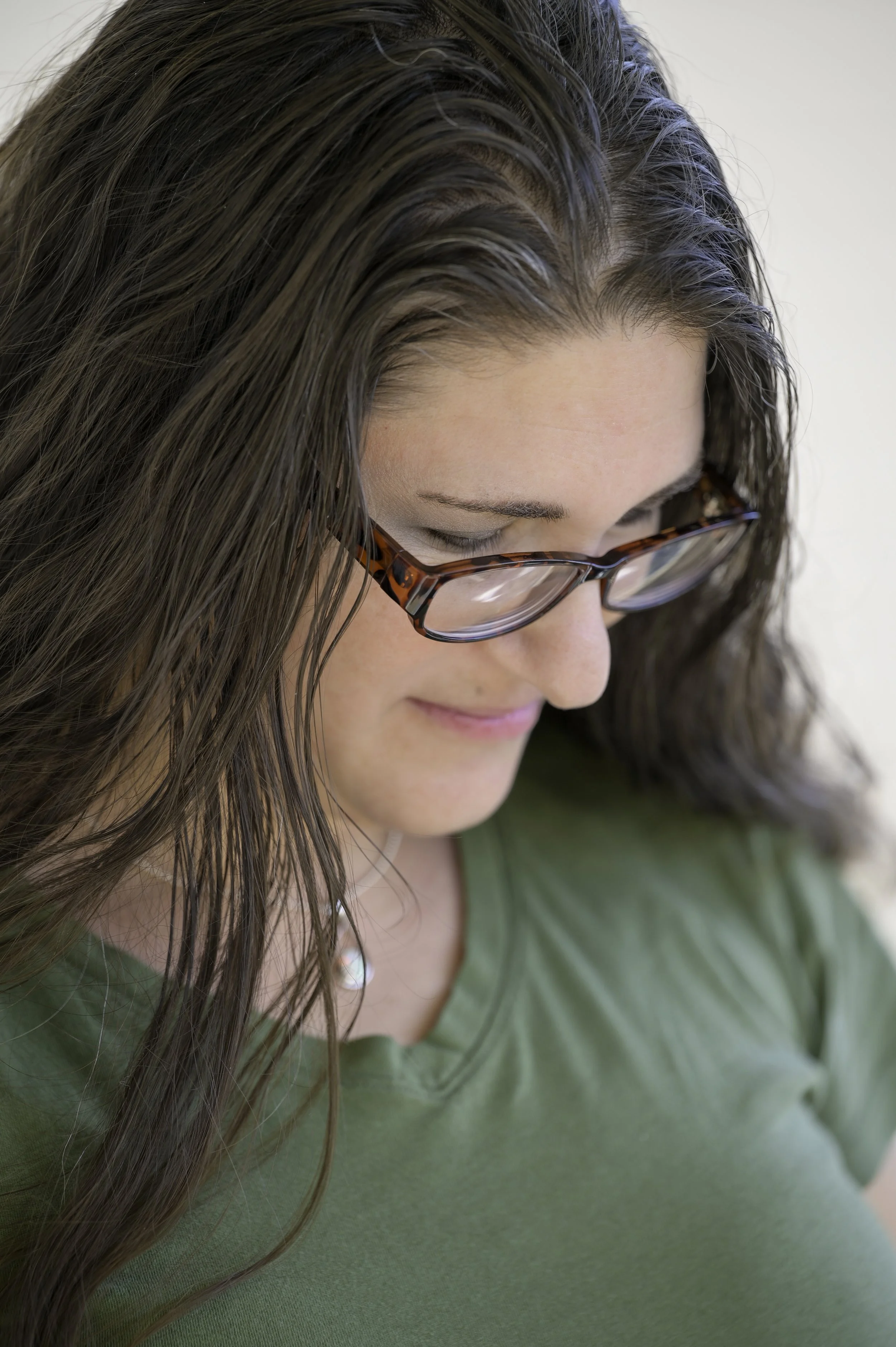 A woman with long brown hair and wearing glasses is looking down and smiling. She is wearing a green top.