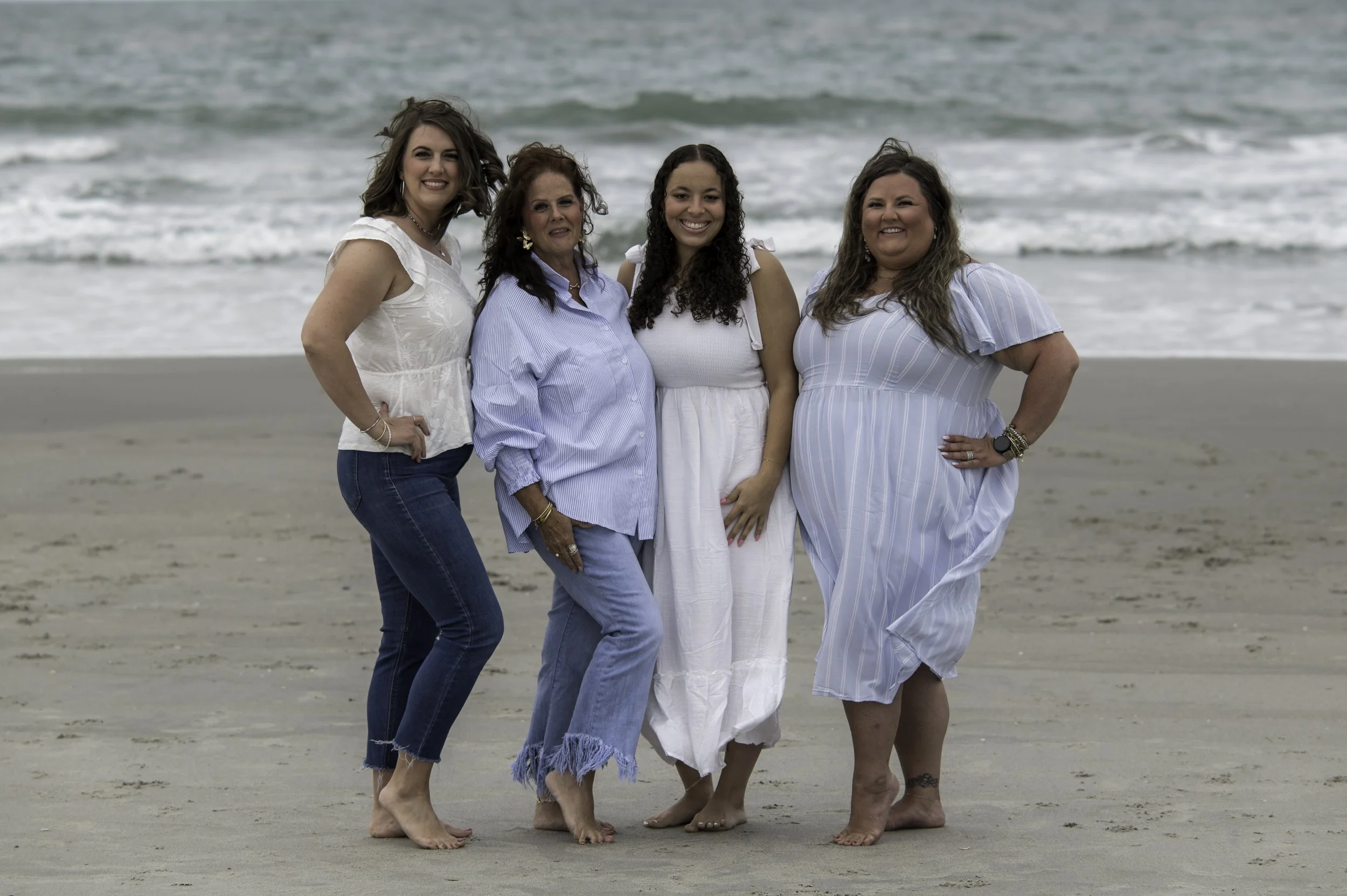 Four women standing together on the beach with ocean waves in the background, smiling at the camera.