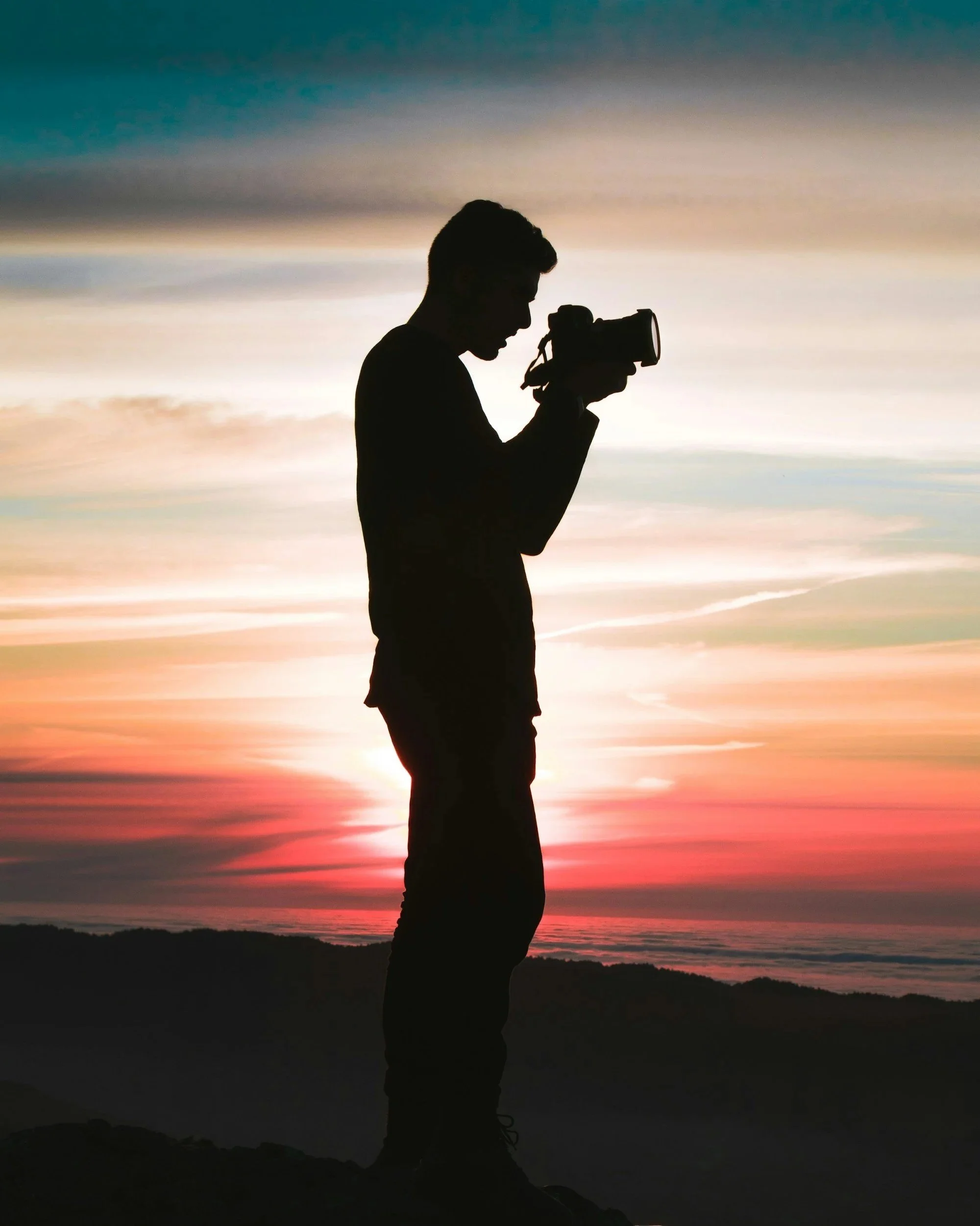 Silhouette of a person taking a photo with a camera during sunset or sunrise over a landscape.