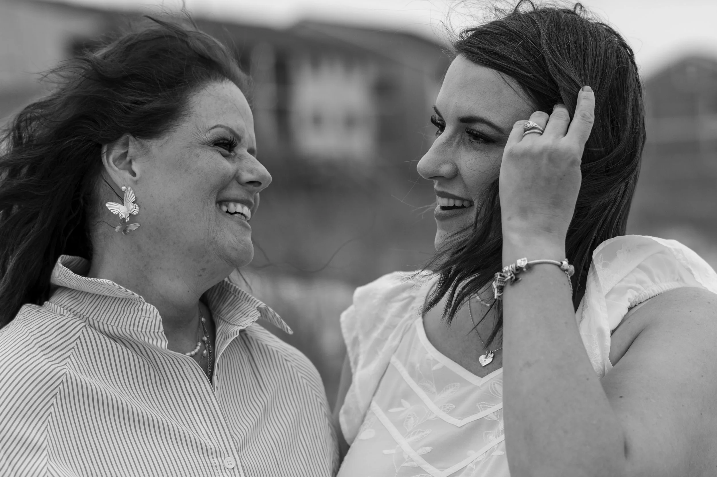 Two women face each other outdoors, smiling and touching each other's hair.