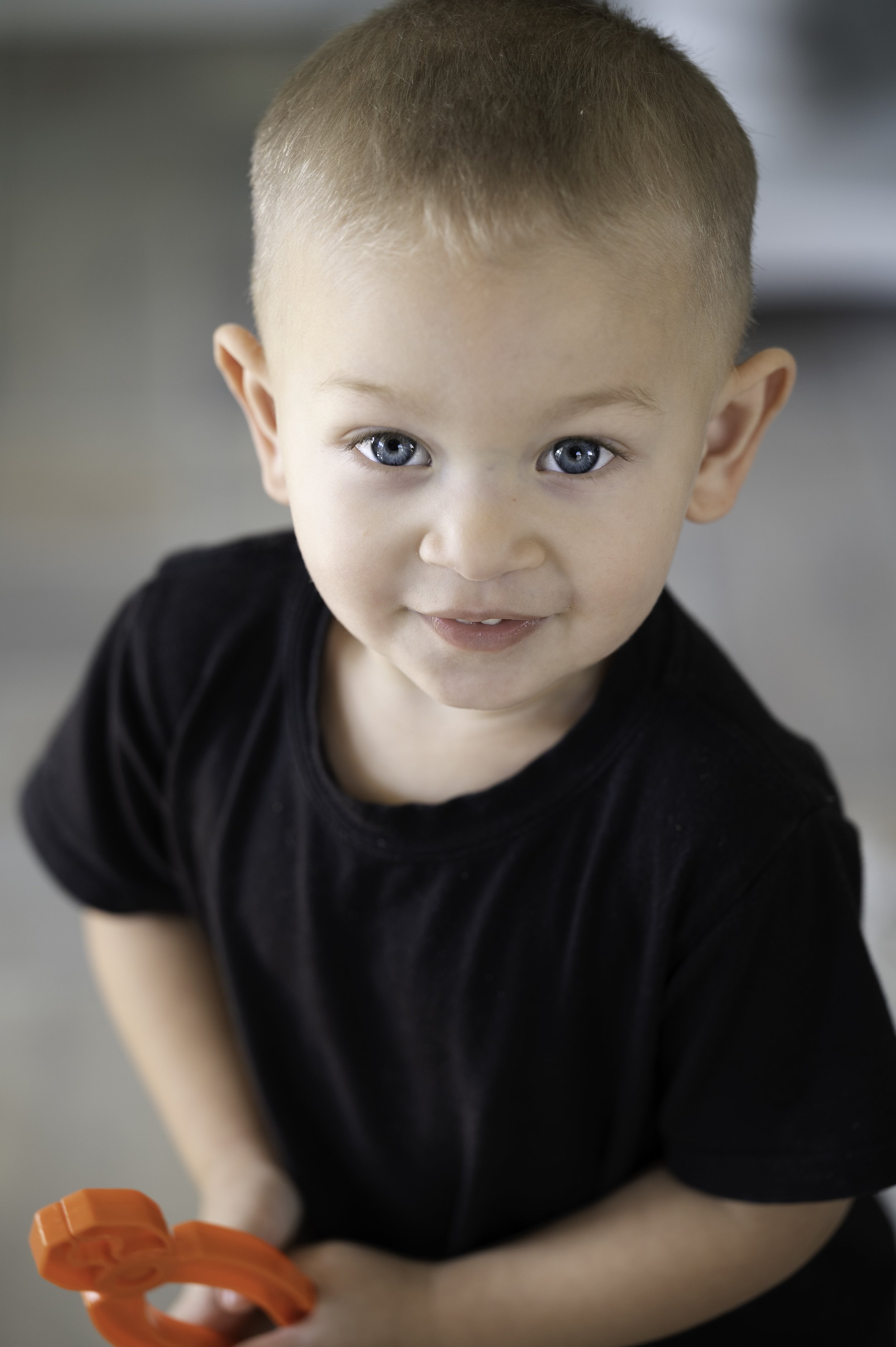 A young boy with blue eyes and short blonde hair, wearing a black shirt, holding an orange toy, and smiling at the camera.