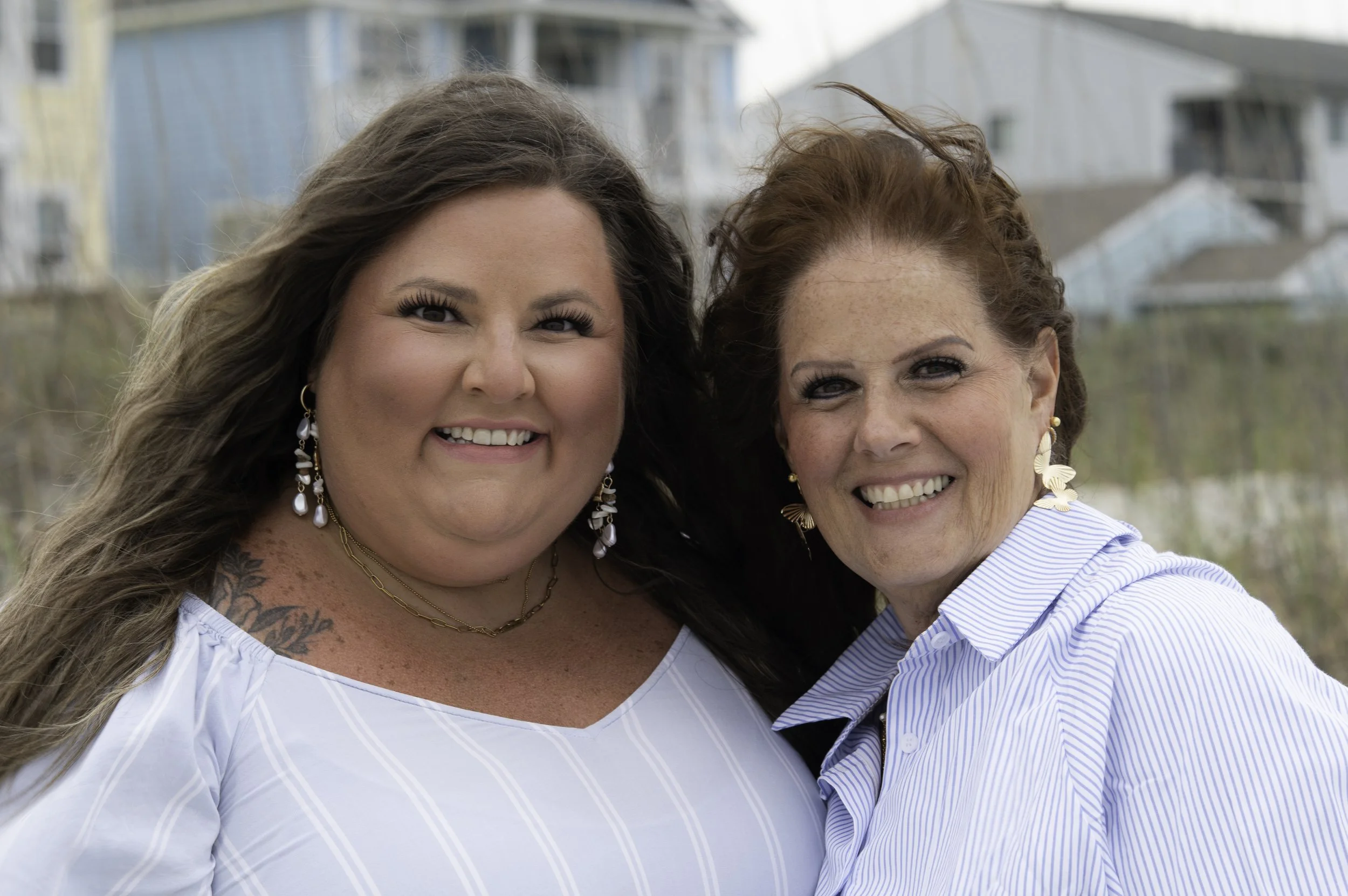 Two women with long hair and earrings smiling outdoors with buildings in the background.