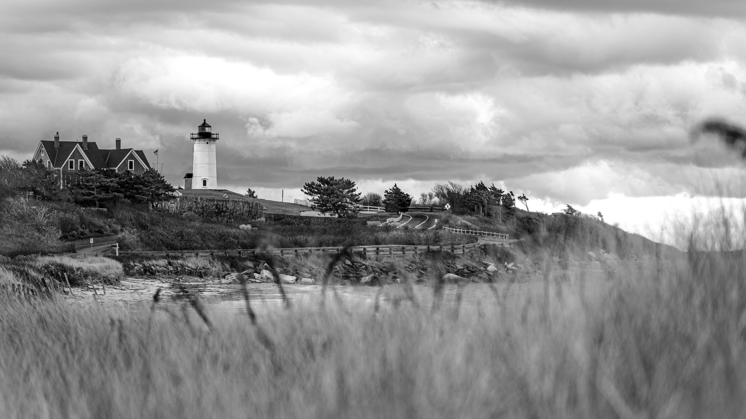 A black and white photo of a coastal scene featuring a lighthouse, a house, trees, a winding road, and tall grass in the foreground, with cloudy skies overhead.