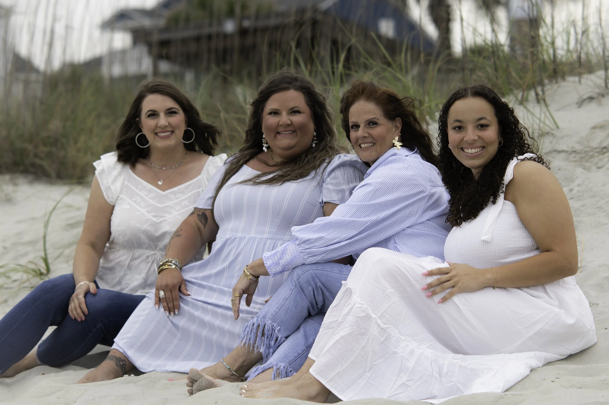Four women sitting together on the beach, smiling at the camera. There is sand and grass in the background with a wooden deck or bridge in the distance.