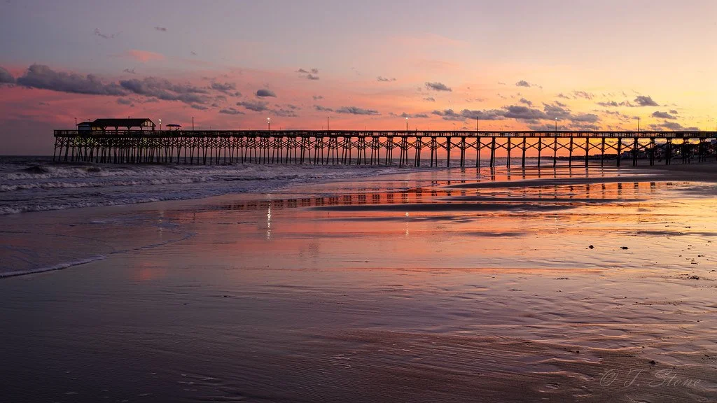 Sunset over a wooden pier extending over the ocean with reflections on wet sand.
