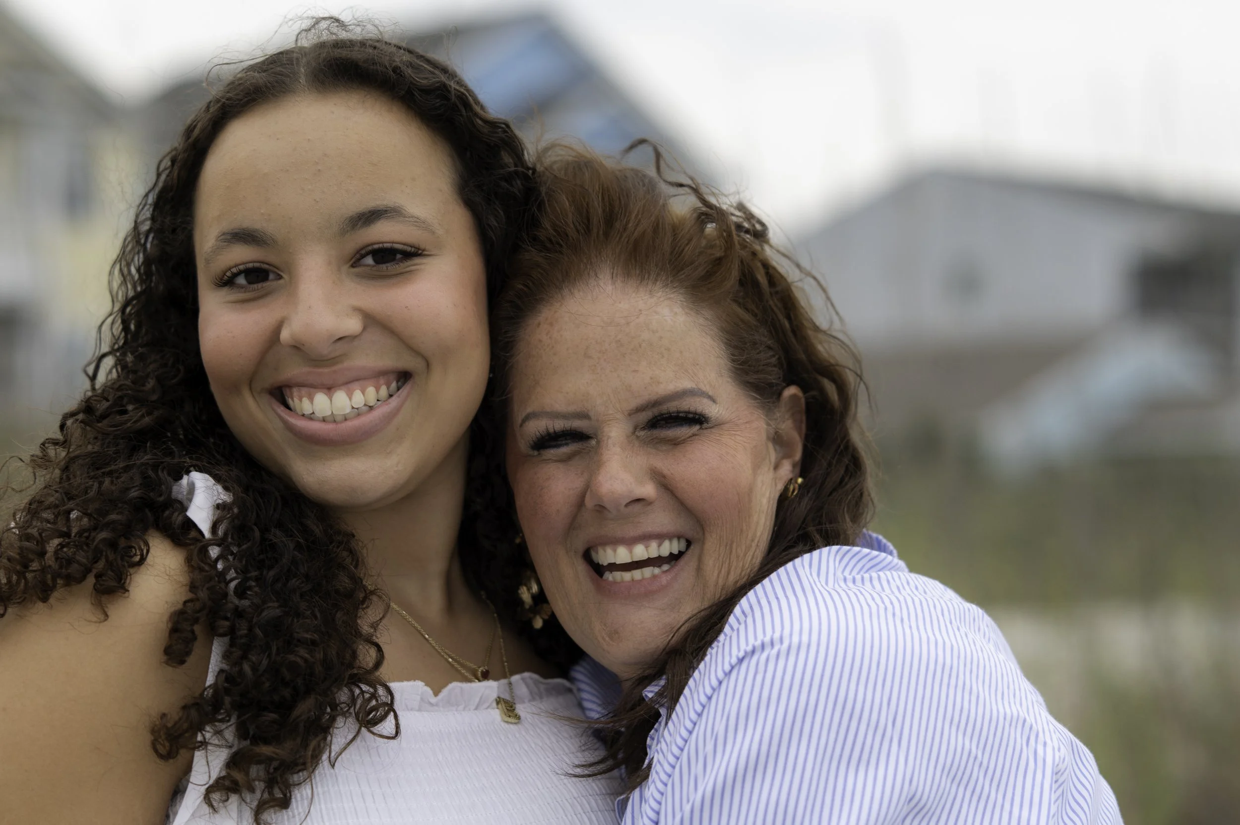 A young woman with curly hair smiling and hugging an older woman with red hair, both outdoors with a blurred background of houses.
