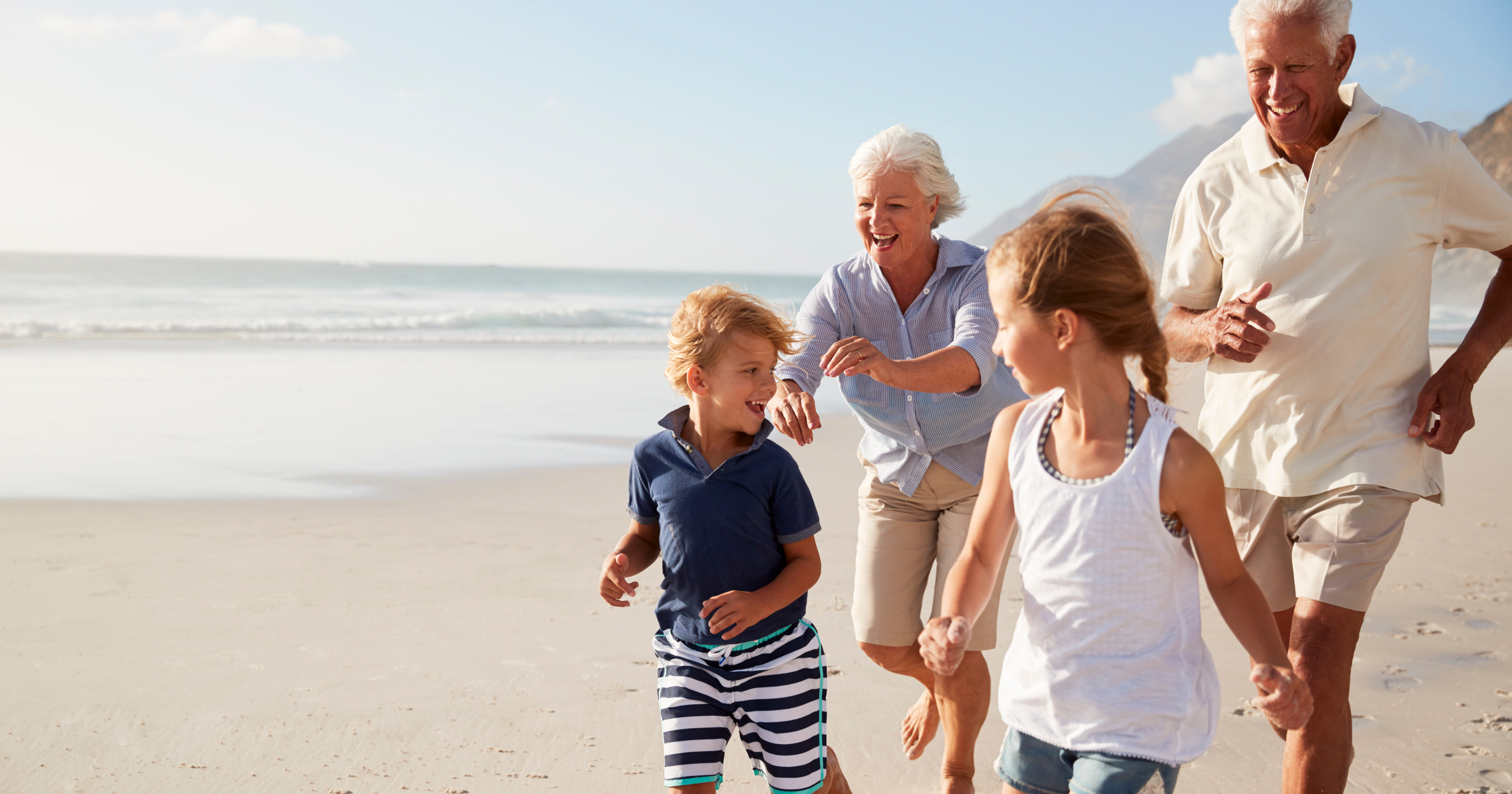 A group of grandparents and children playing and running on the beach with the ocean and mountains in the background on a sunny day.