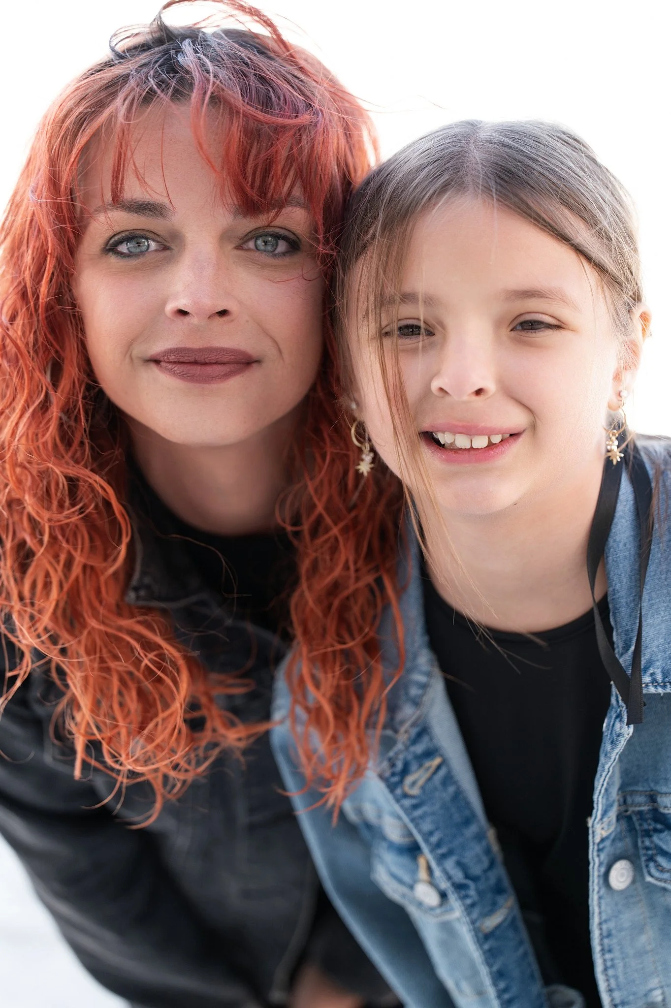 A woman with red curly hair and a young girl with light brown hair, both smiling, close together, wearing casual clothing, outdoors in a bright setting.
