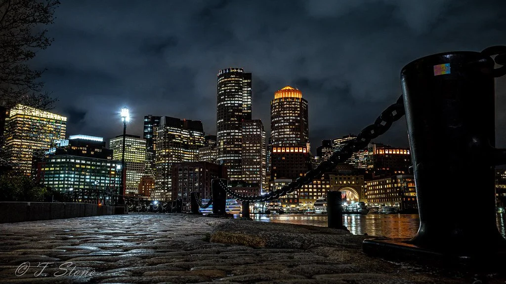 Nighttime view of a city skyline with tall buildings illuminated, seen from a cobblestone waterfront promenade with a chain barrier in the foreground.