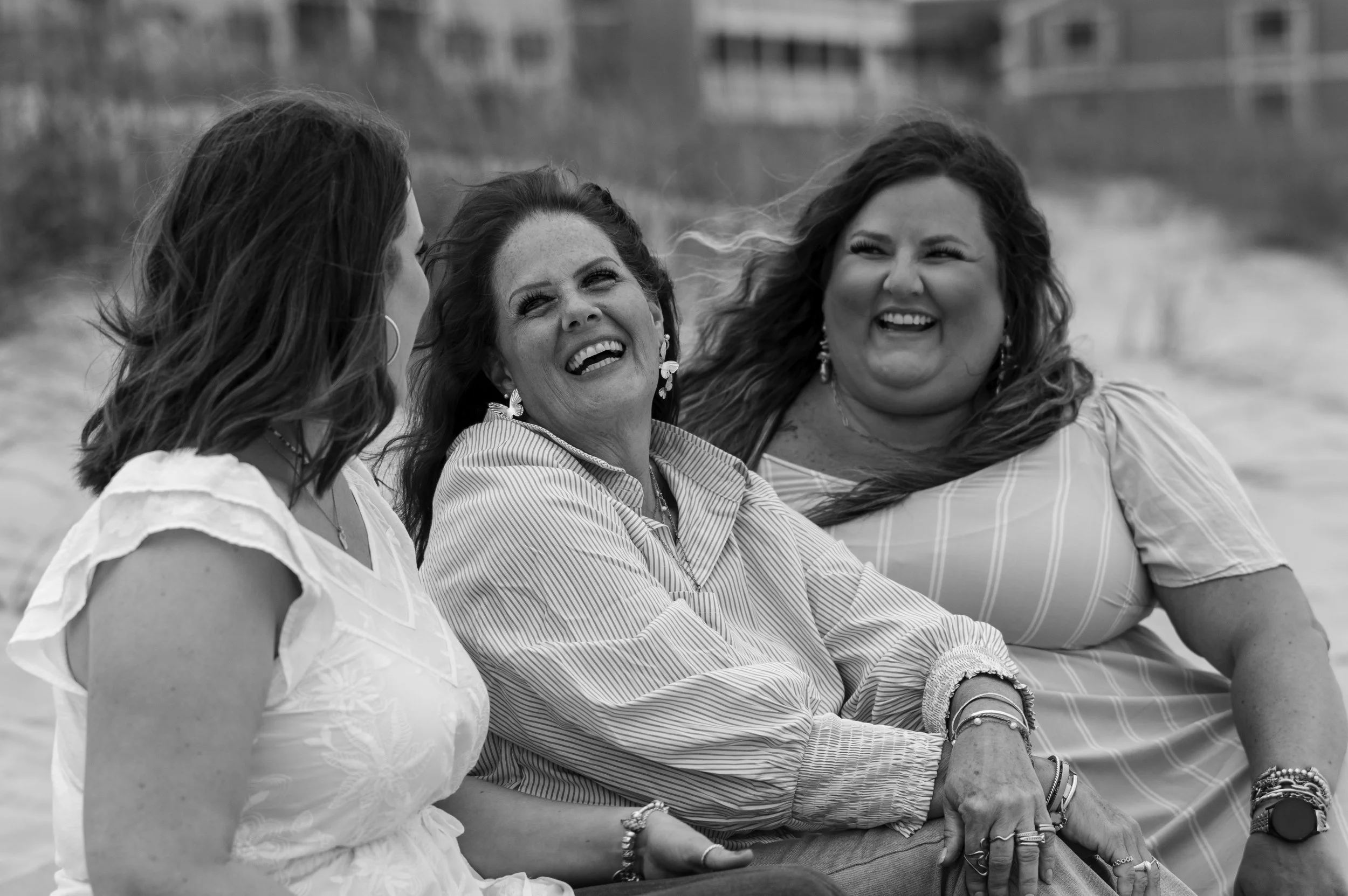 Three women sitting outdoors, smiling and engaging in conversation, with a blurred background of a body of water and buildings.