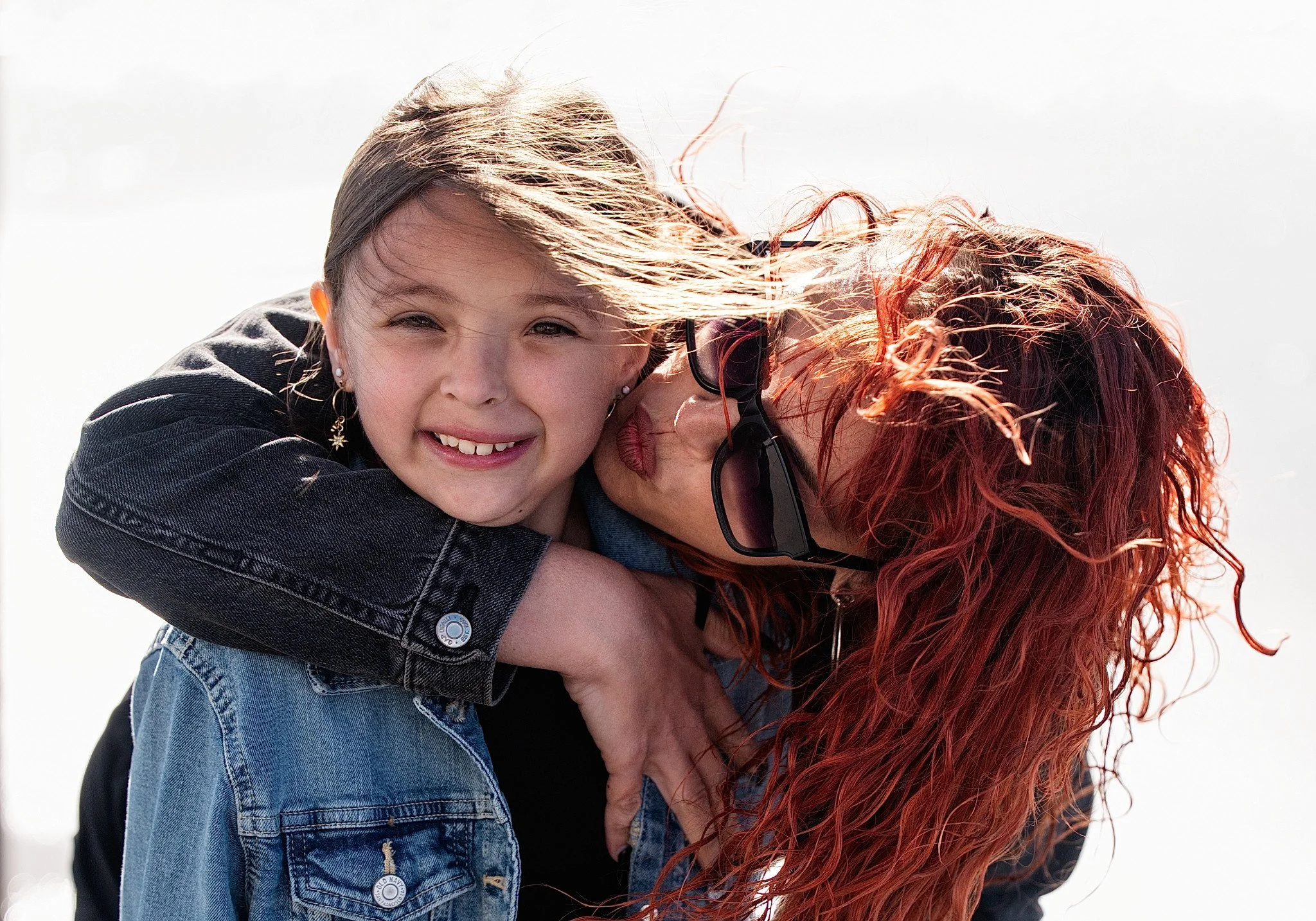 A woman and a girl are outside, with the woman hugging the girl from behind. The girl has light brown hair, is smiling, and is wearing earrings and a denim jacket. The woman has red curly hair, sunglasses, and is leaning in close to the girl.